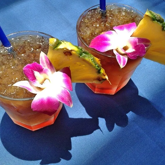 Two Mai Tai cocktails garnished with pineapple slices and orchids on a blue tablecloth.
