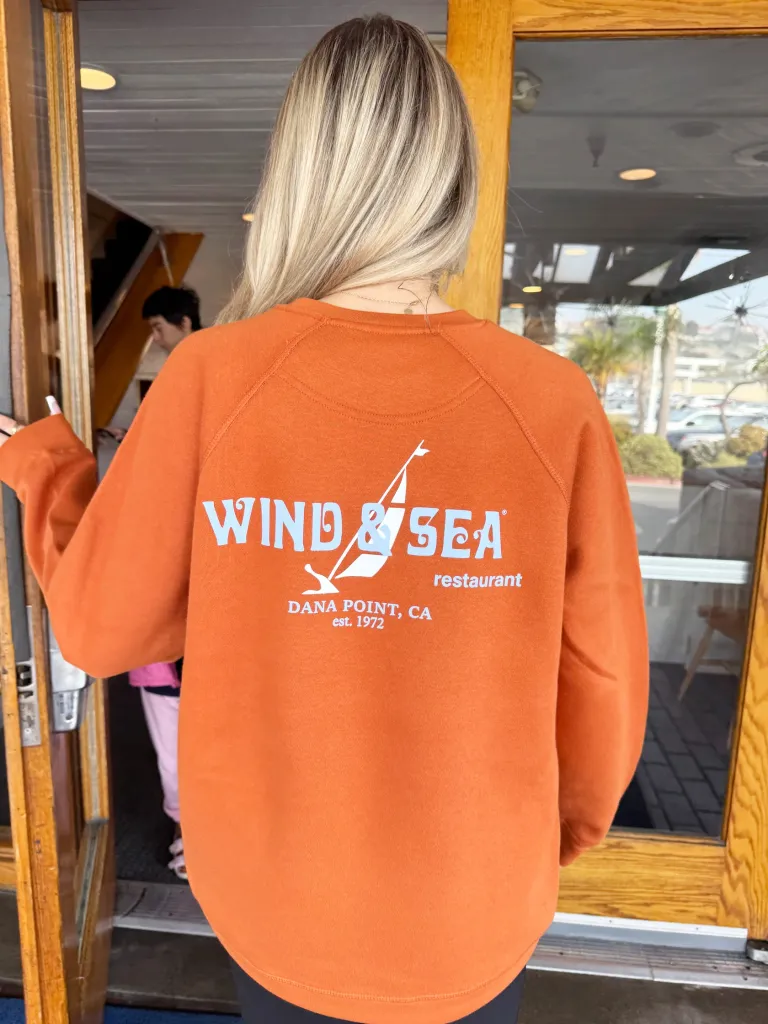 A blonde person from behind wearing an orange Wind & Sea Restaurant sweatshirt.