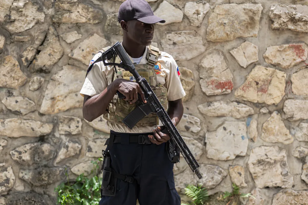 A man in a military-style uniform and tactical vest holds a rifle while standing in front of a stone wall in Port-au-Prince, Haiti.