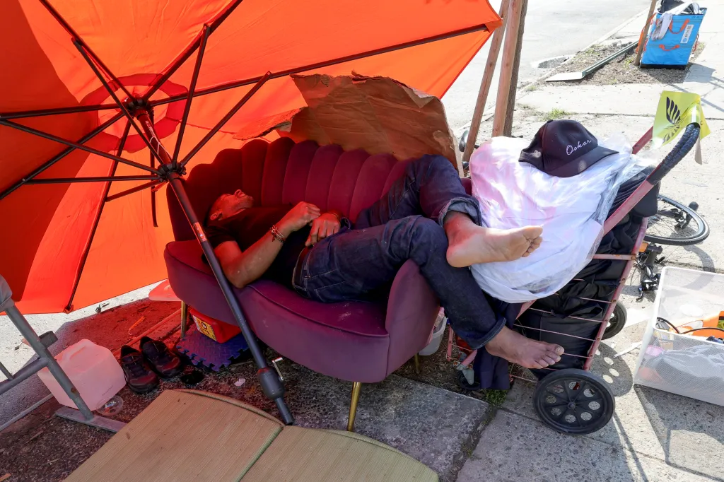 A homeless man sleeps on a couch under an orange beach umbrella on a sidewalk.