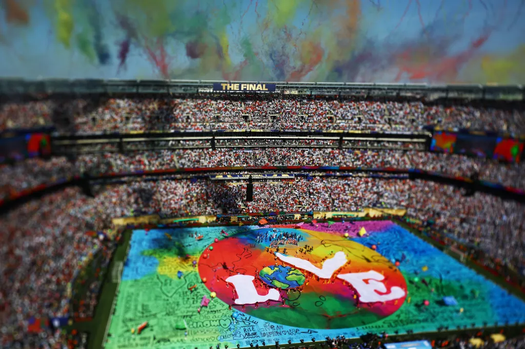 A high-angle view of a stadium during a halftime show, with a large, colorful display on the field and smoke rising into the air.
