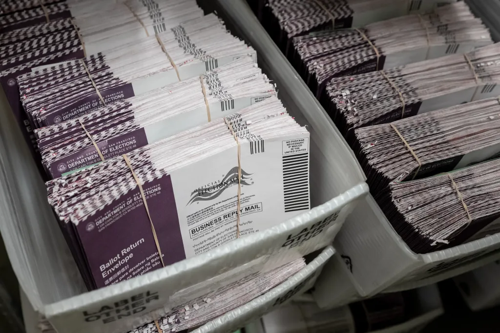 Stacks of mail-in ballot return envelopes, banded together with rubber bands, in a white bin at an election facility.