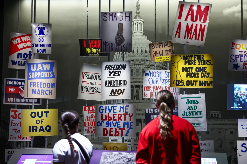 Two people read protest signs in the American Democracy exhibit at the Smithsonian American History Museum, with signs advocating for D.C. Statehood, 