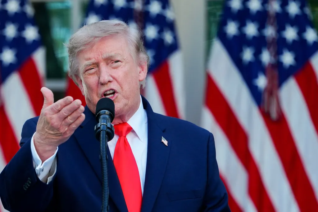 Donald Trump speaking into a microphone with two American flags in the background.