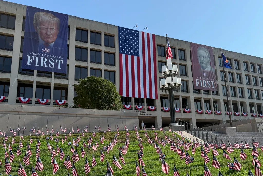 Banners of Donald Trump, an American flag, and another political figure hang from a building, with small American flags planted in the grass in front.