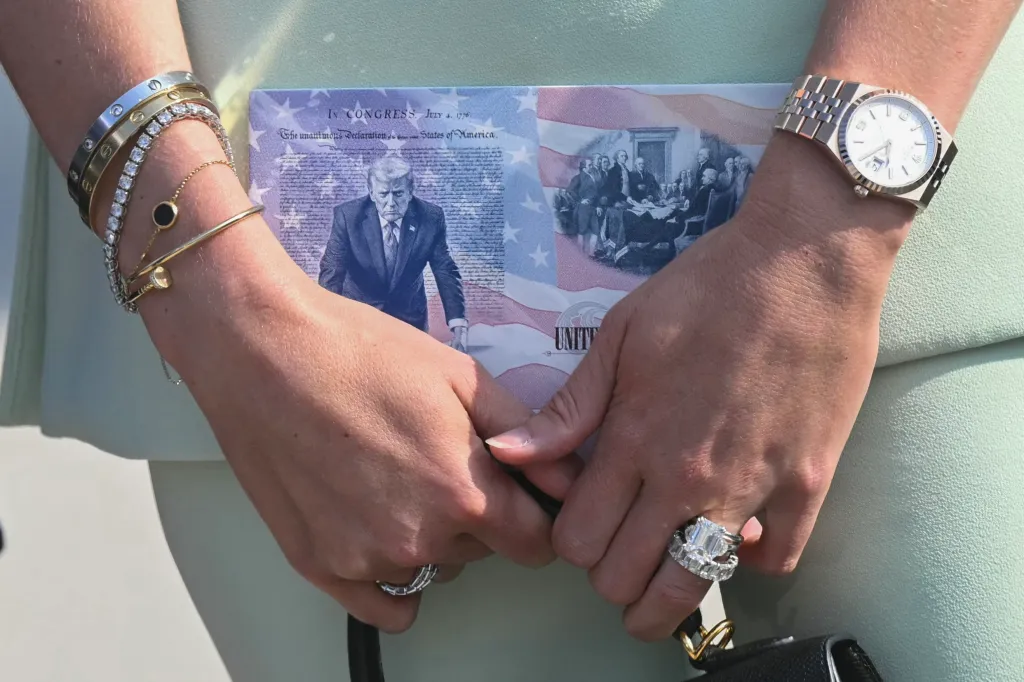 A woman's hands, adorned with rings and bracelets, hold a document featuring an image of Donald Trump, the Declaration of Independence, and a historical depiction of American founding fathers.