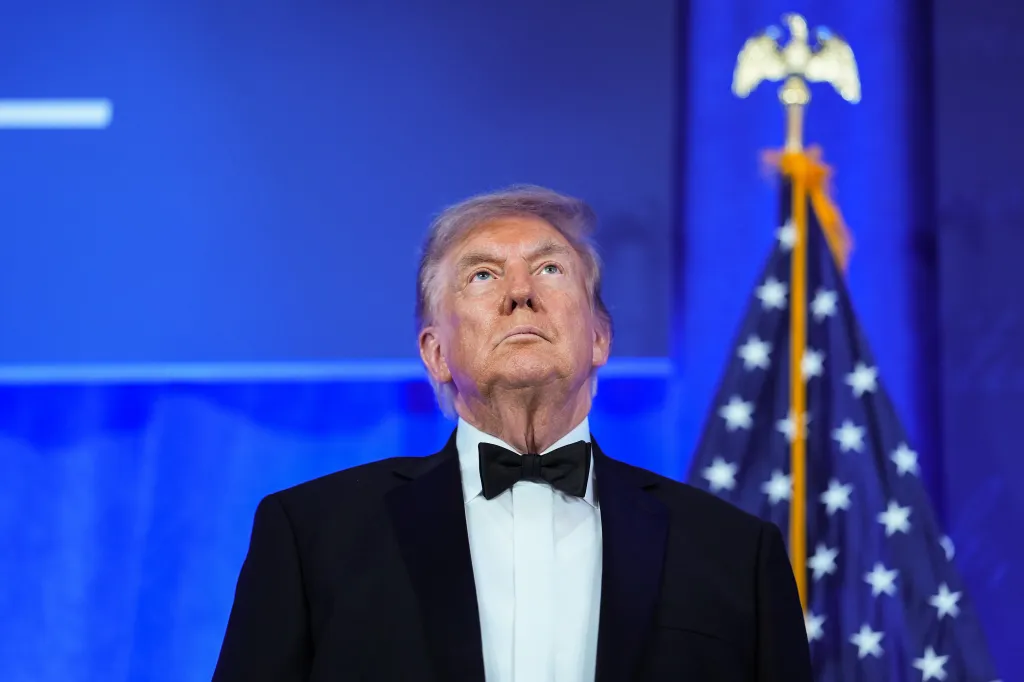 President Trump, in a black tuxedo and bow tie, looks upwards with an American flag to his right, against a blue background.