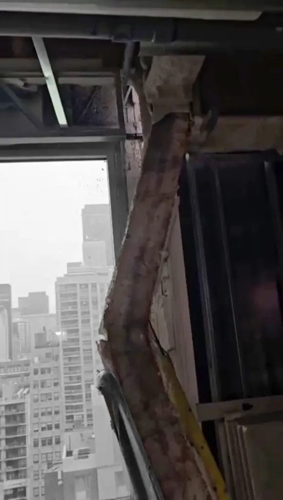 A buckled support column inside a building, with city skyscrapers visible through a window.