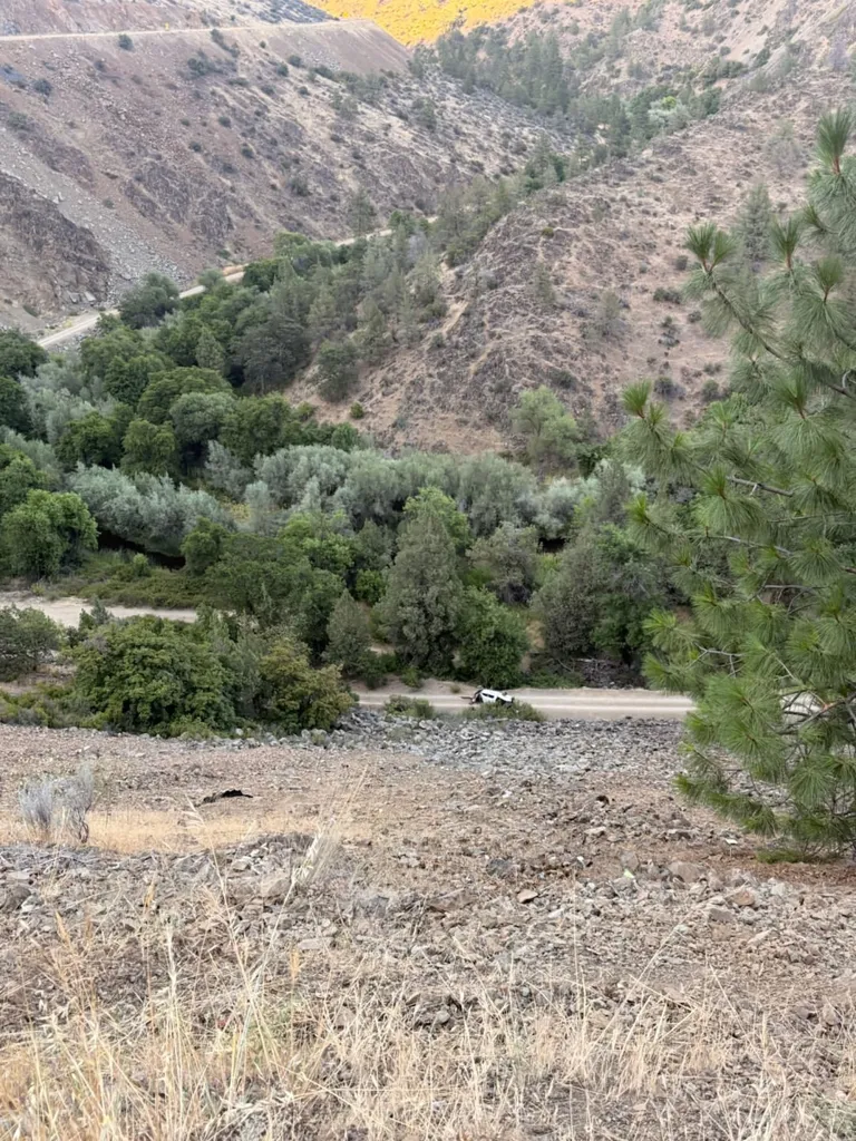 A white truck lies overturned at the bottom of a steep, rocky cliff, having plunged off the highway visible at the top.