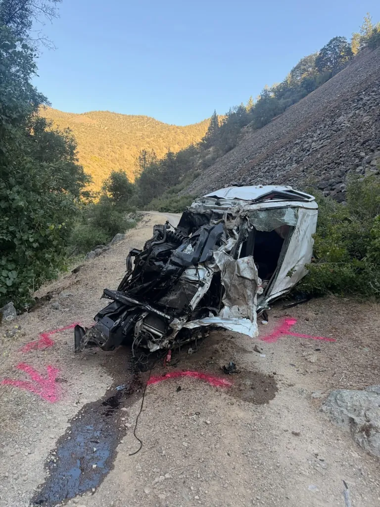 A wrecked white pickup truck with its engine severely damaged lies on a dirt road, marked with pink spray paint.
