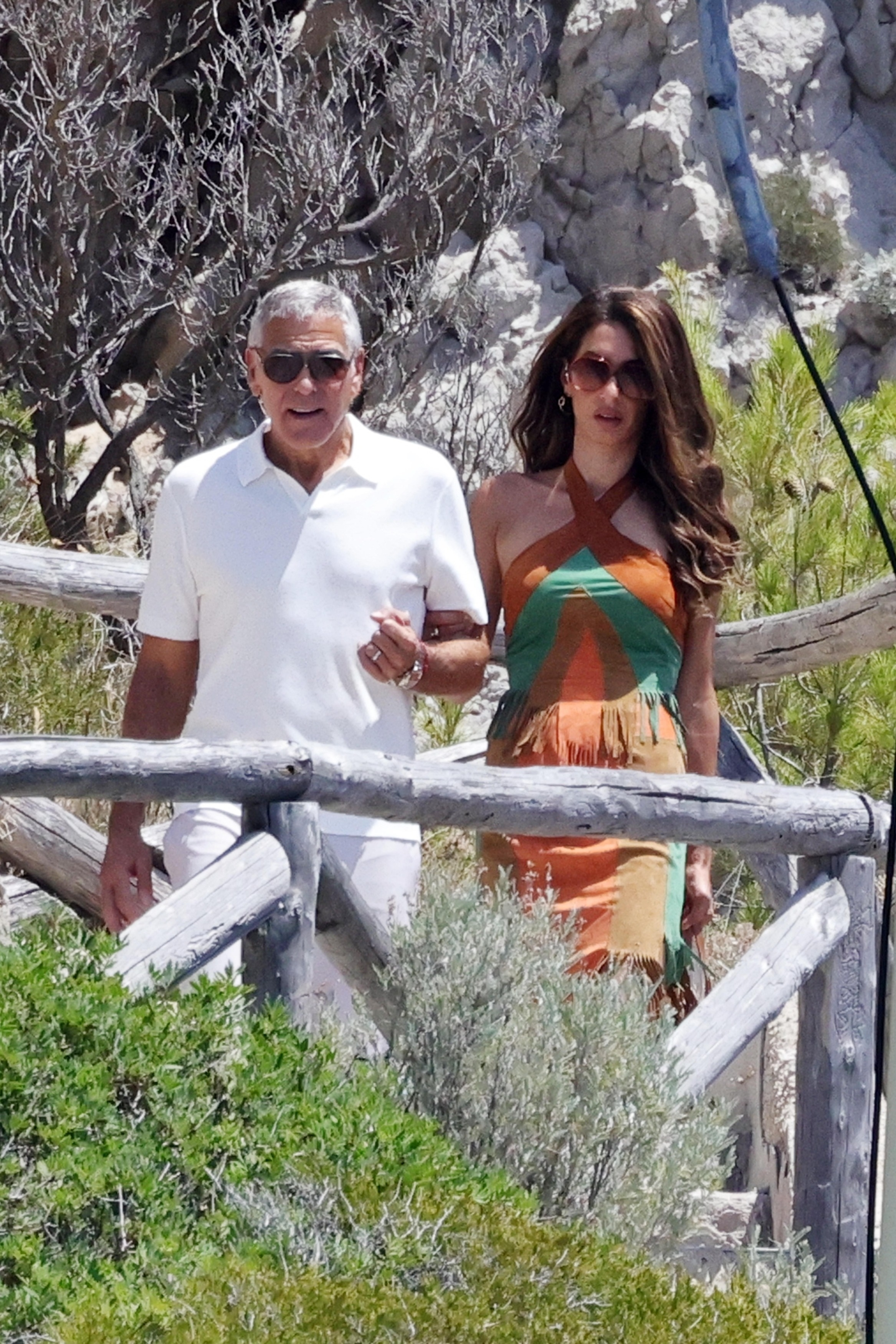 George and Amal Clooney walk arm-in-arm on a path surrounded by lush greenery, with George in a white polo and Amal in a fringed halter dress.
