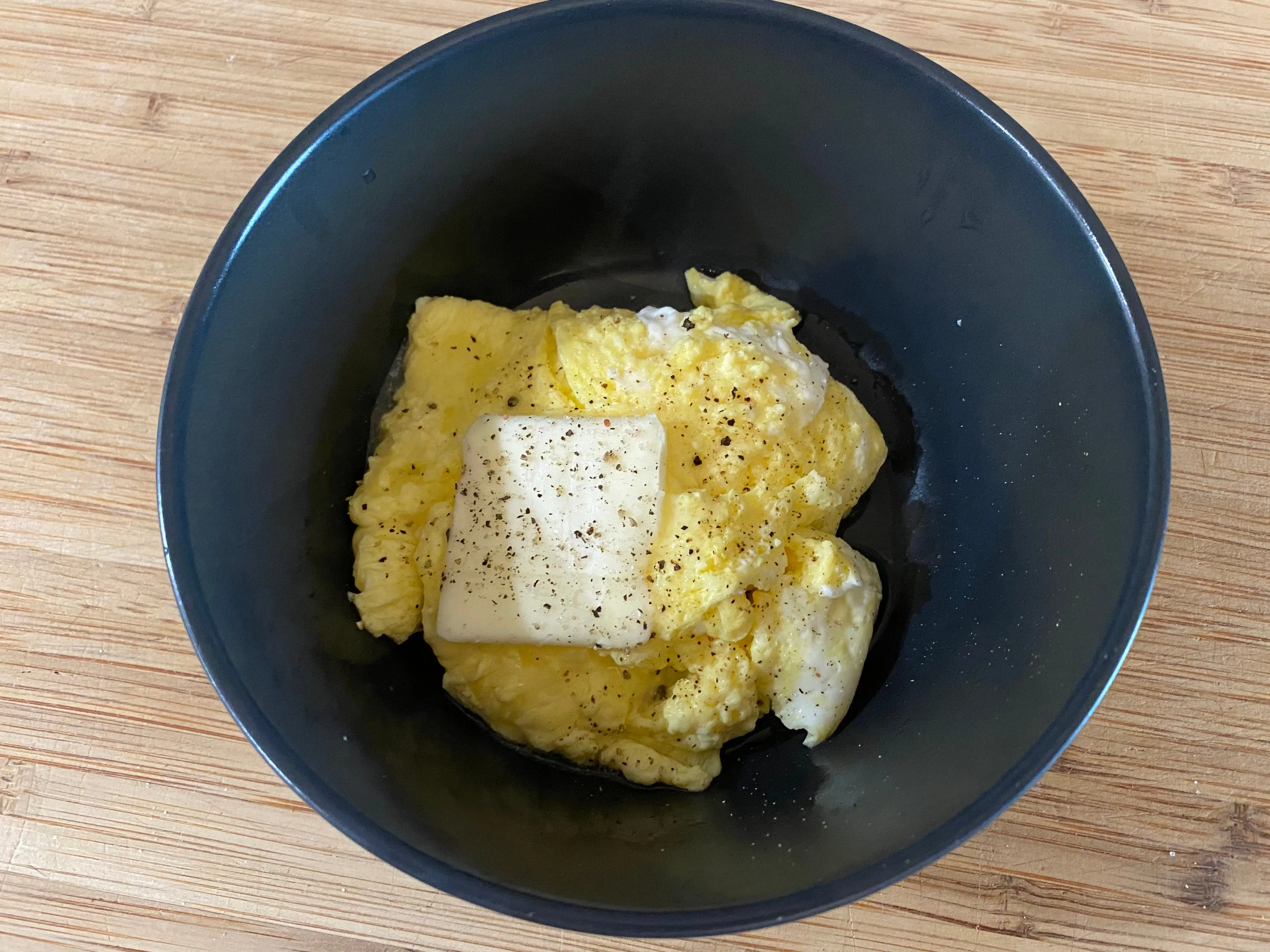 Scrambled eggs in a blue bowl with pepper and a pad of butter sit on a wooden table
