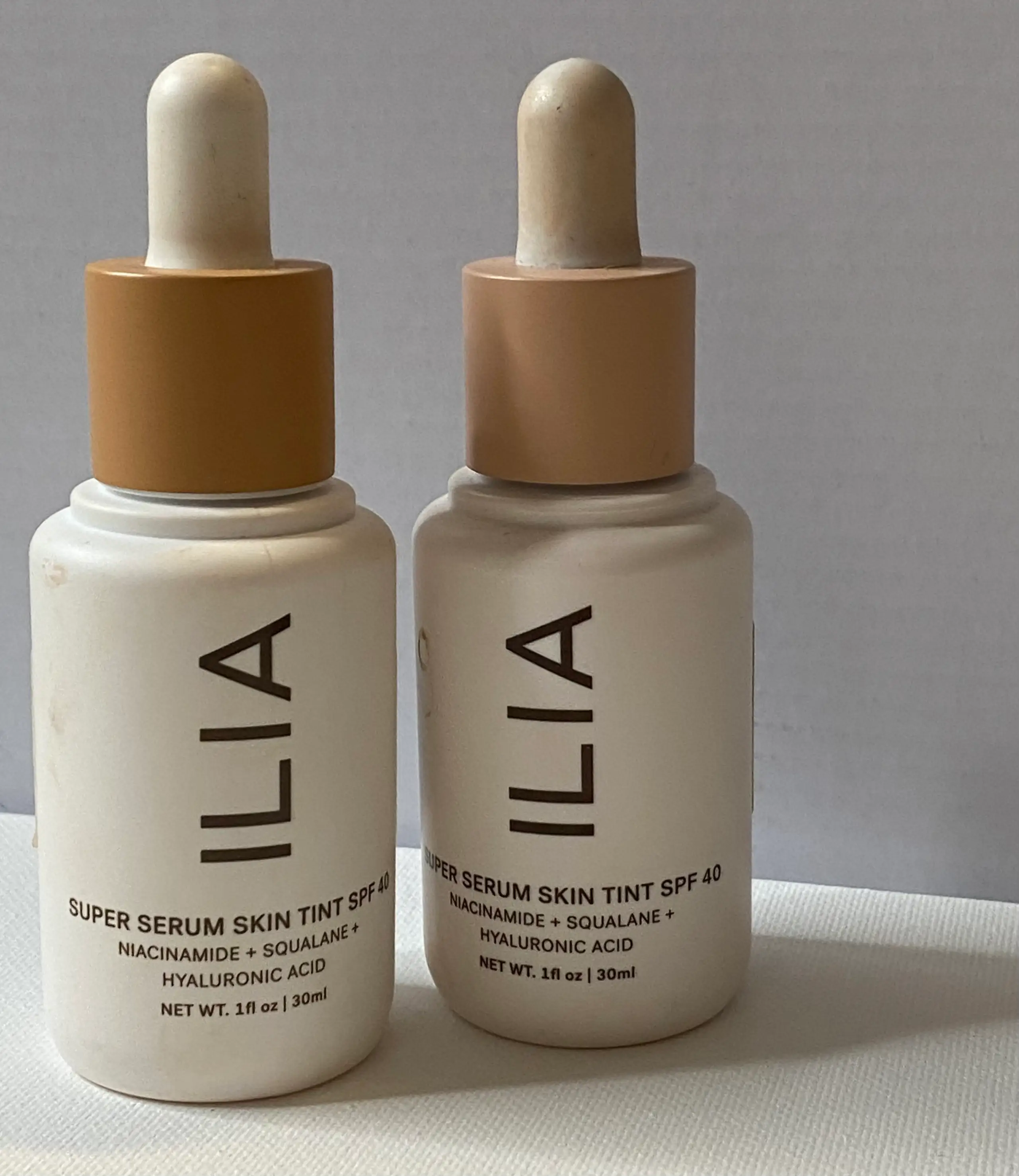 Two white bottles of ILIA skin tint with white dropper and tan bands around the top against a white background