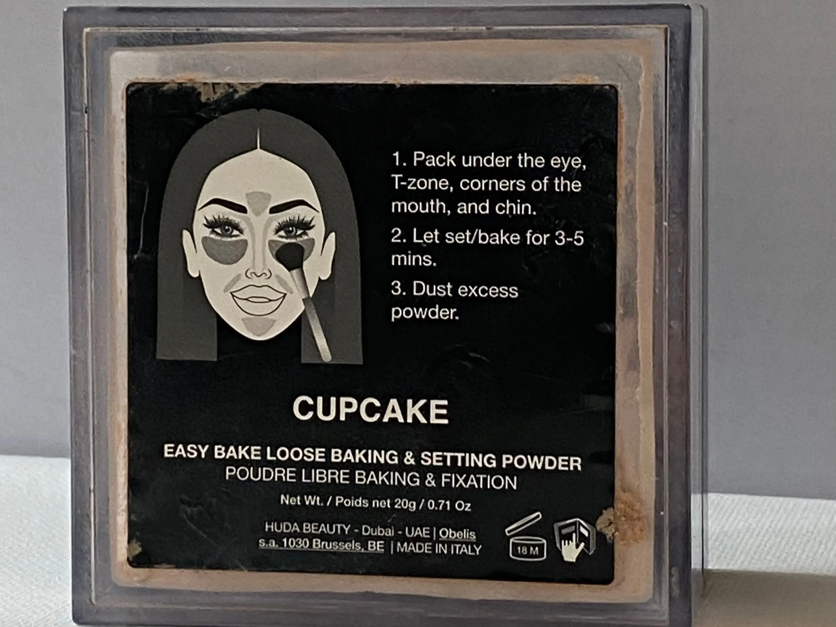A loose powder box with an illustration of a woman with a makeup brush and a diagram of where to place product with directions on black label