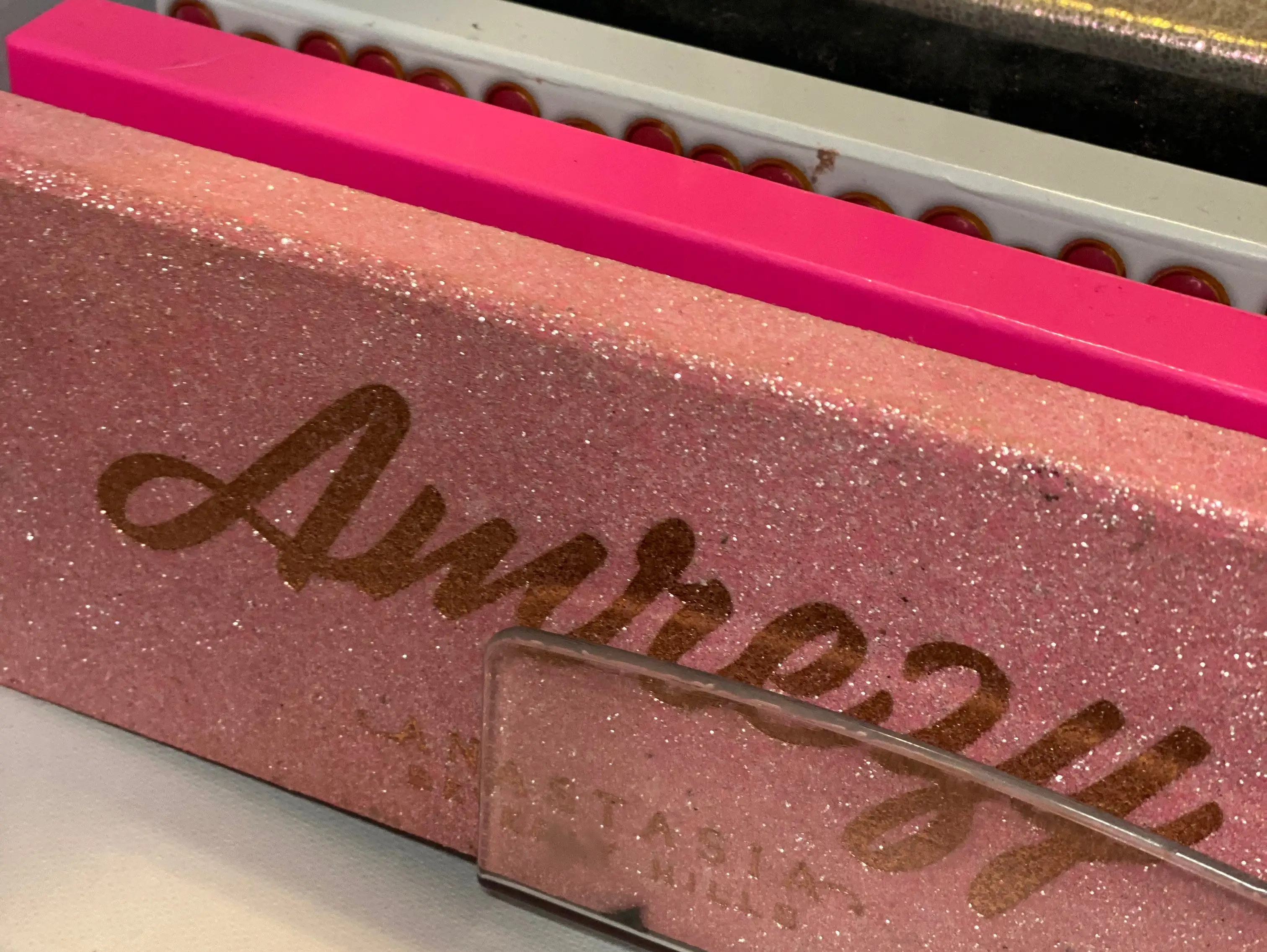 Collection of eyeshadow palettes in a drawer with only spines visible except front palette. Front palette is pink and sparkly with gold text that says 
