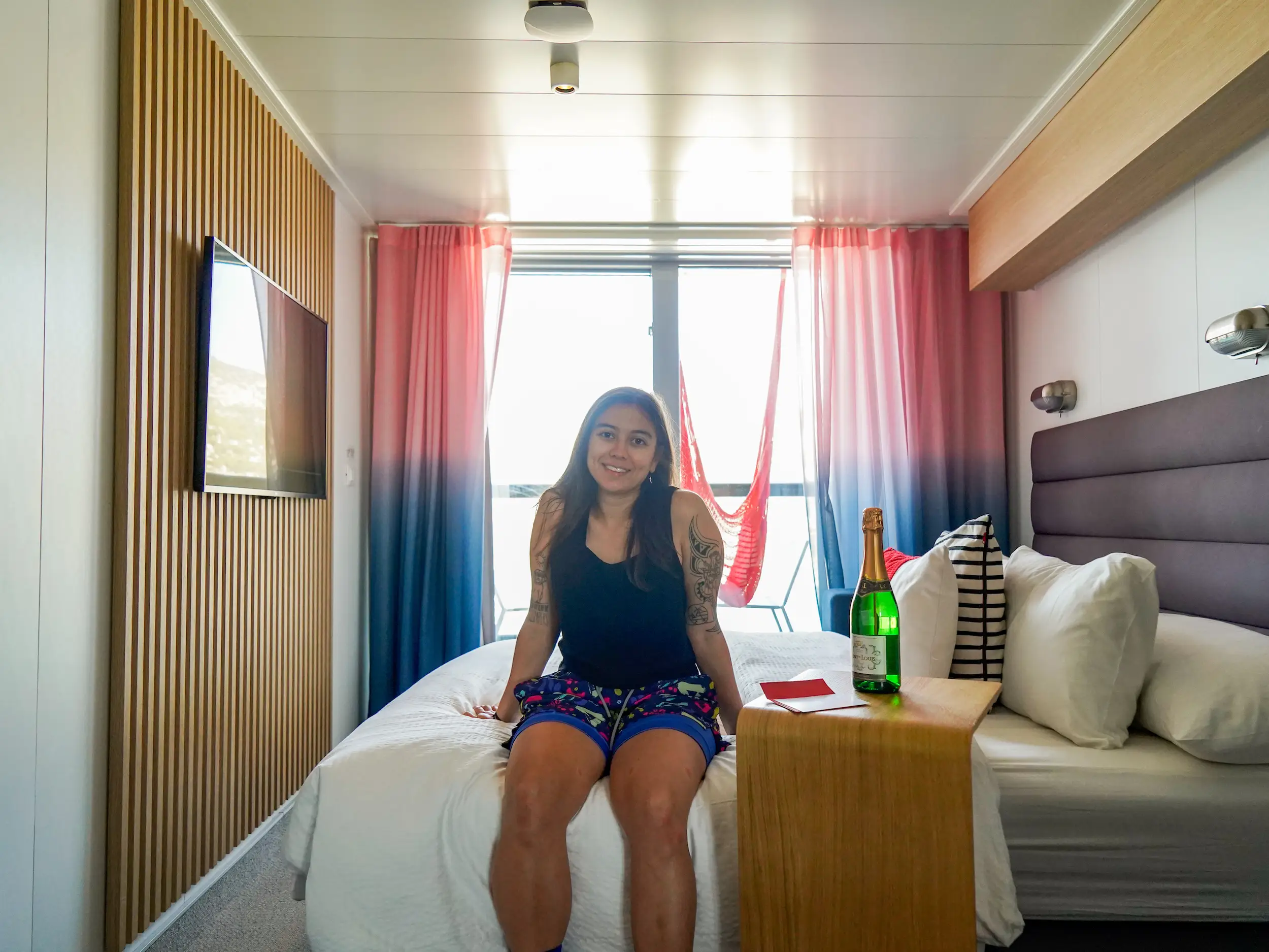 The author sits on a white bed with a TV mounted on the left and open red and blue curtains on the floor-to-ceiling window behind the bed.