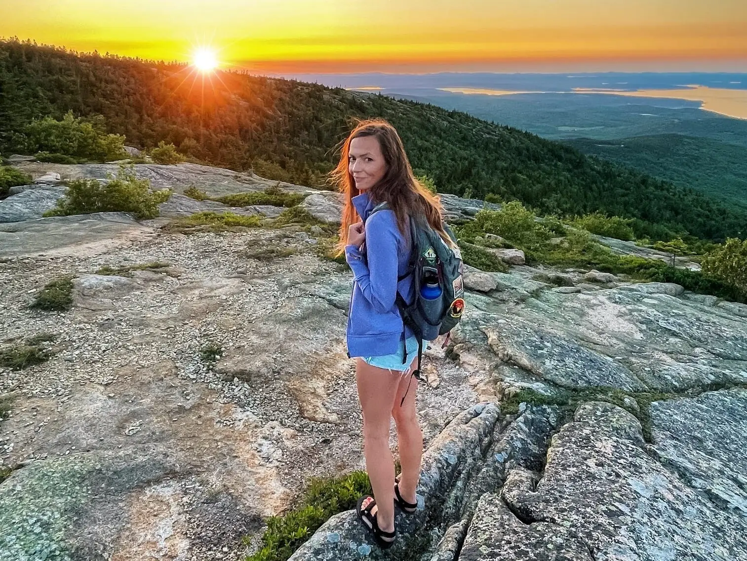 A woman with a blue sweatshirt, shorts, and a backpack at Acadia National Park at sunset.