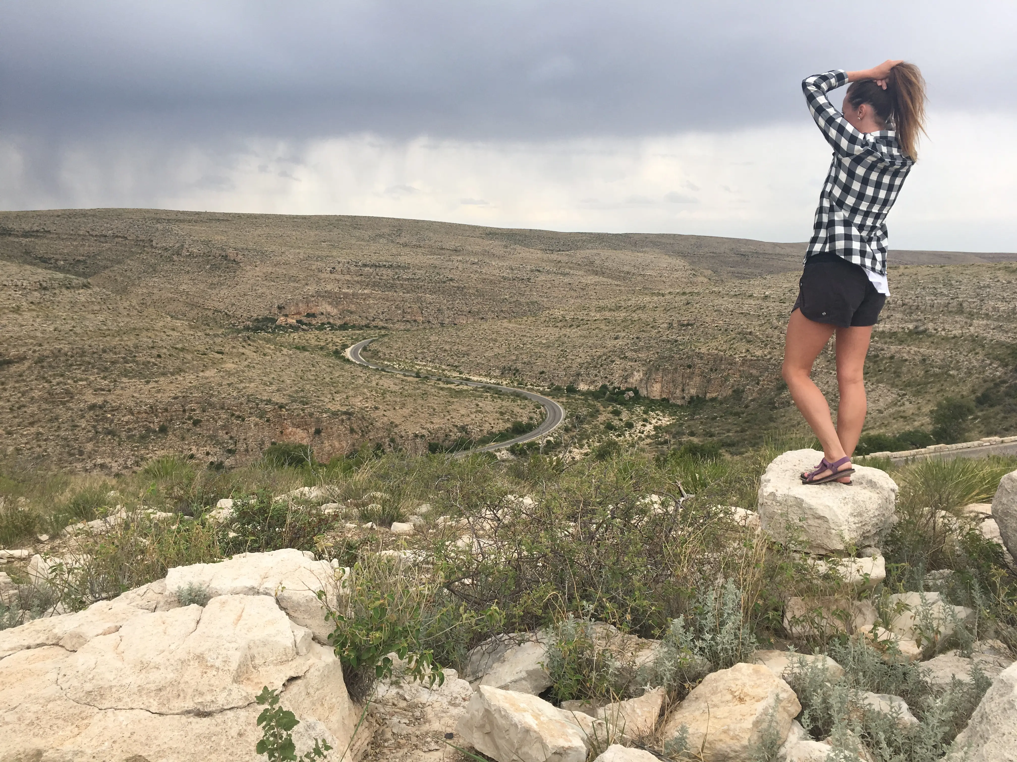 Emily, wearing a black-and-white checkered shirt and black shorts, stands on a rock and puts her hair in a ponytail while looking out at the mountains.