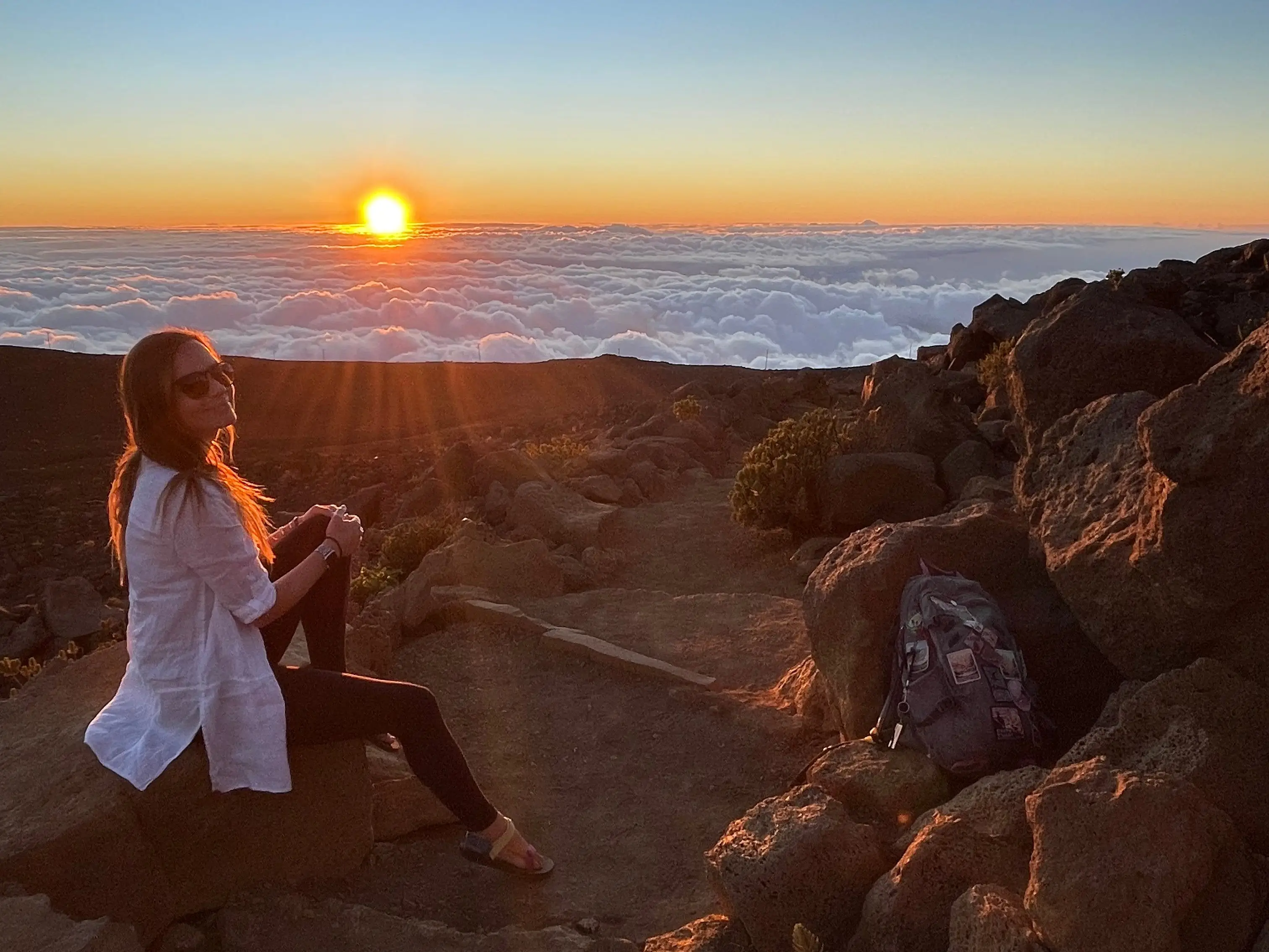 Emily sits on a rock and smiles at the camera, while the sun sets on the beach behind her.