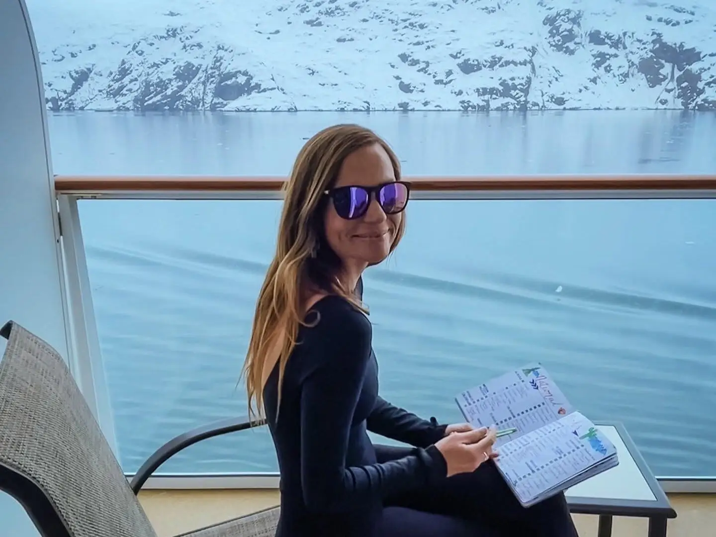 Emily, wearing sunglasses, smiles on a cruise deck that's passing a glacier, as she sits in a chair and writes in a notebook.