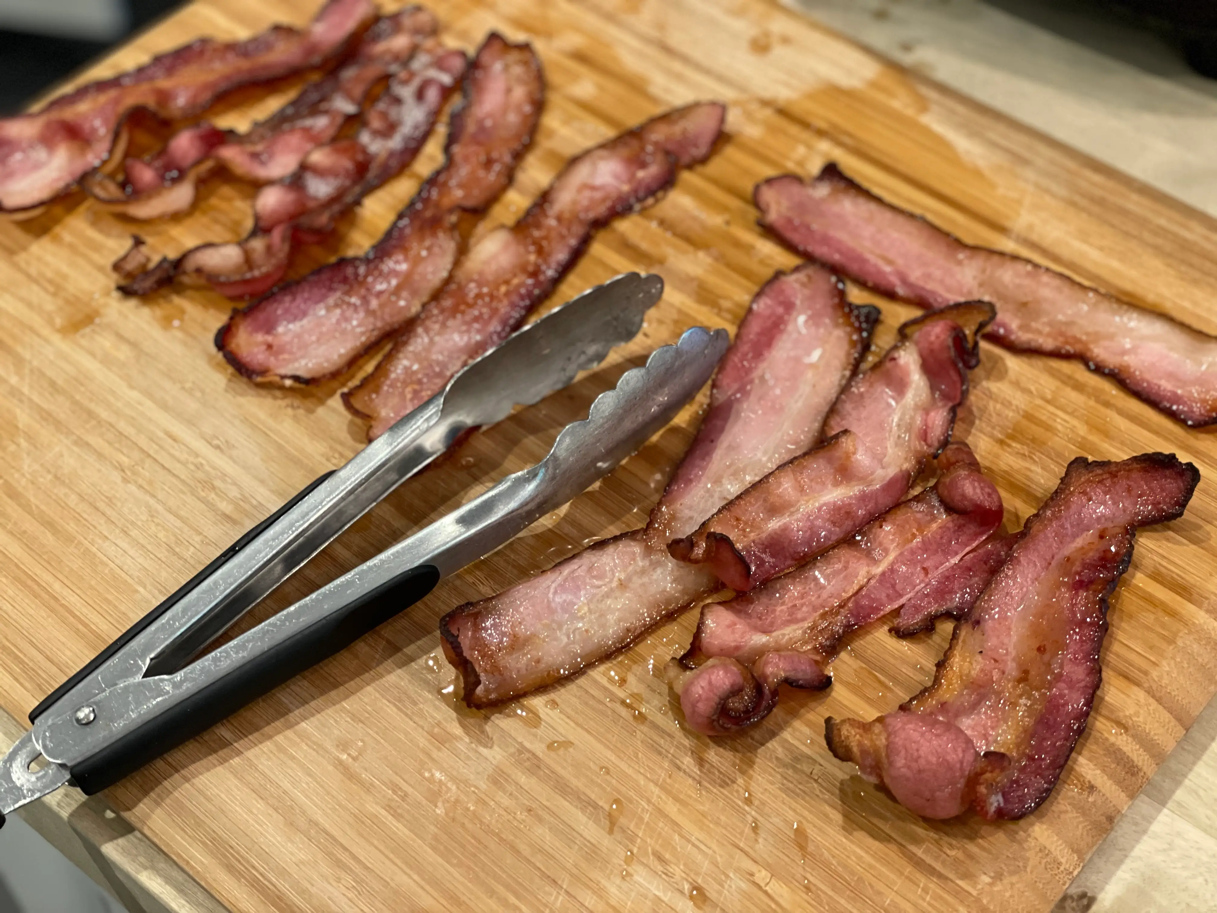 strips of cooked bacon on a cutting board with a pair of metal tongs
