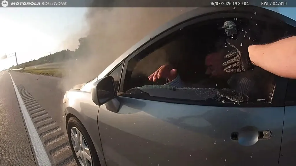 A police officer in a tactical glove breaks the passenger window of a smoke-filled car.