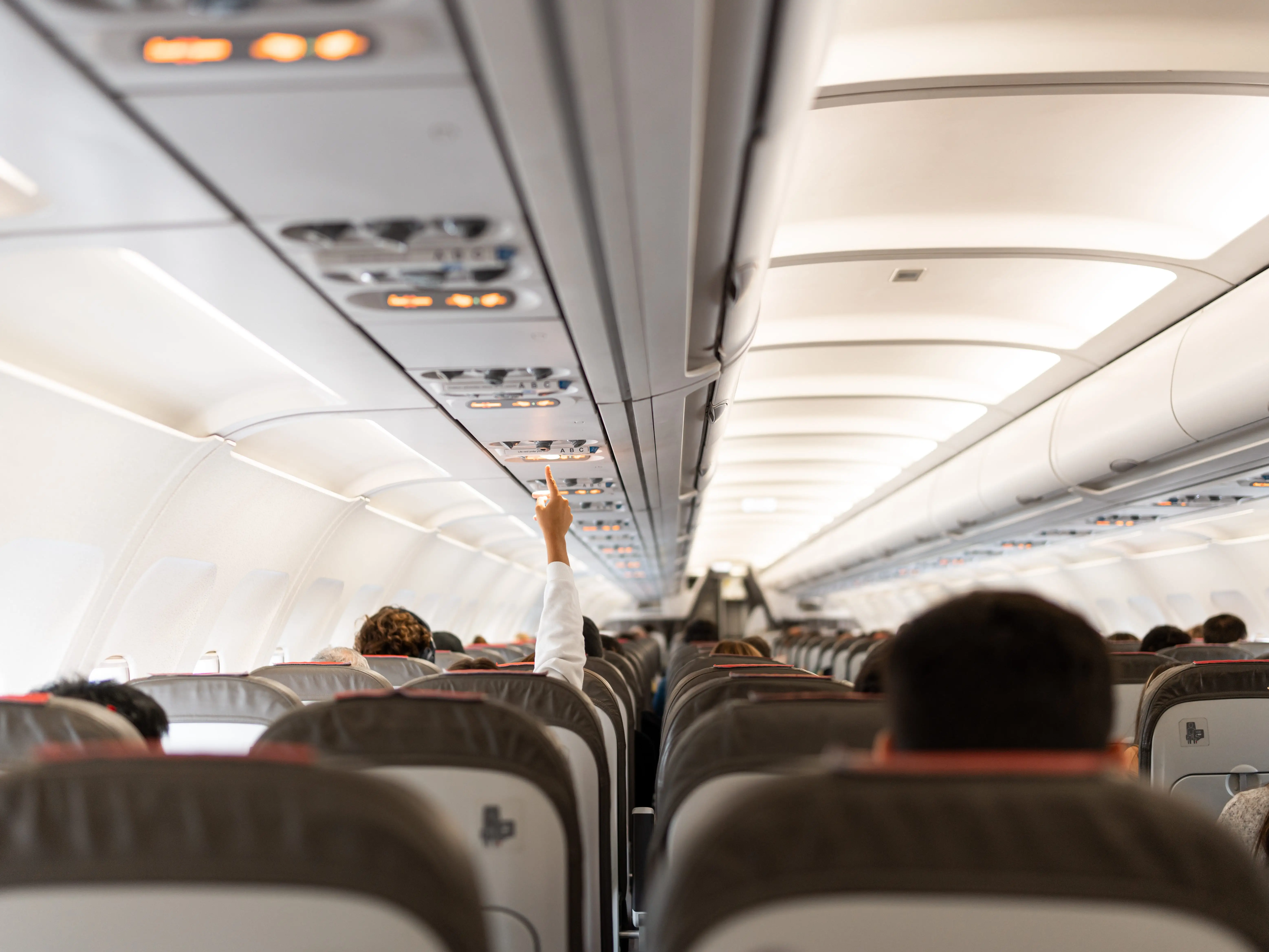 People sitting on rows of seats on a plane, with one person reaching their hand up to call for a flight attendant.