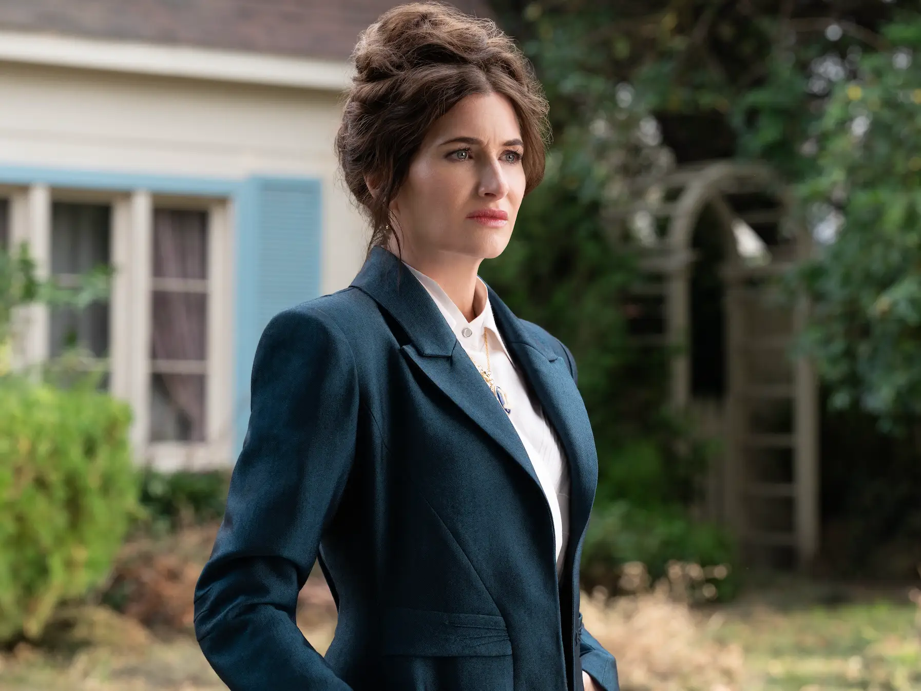 A woman with brown hair in a blue coat and white shirt standing in a garden.