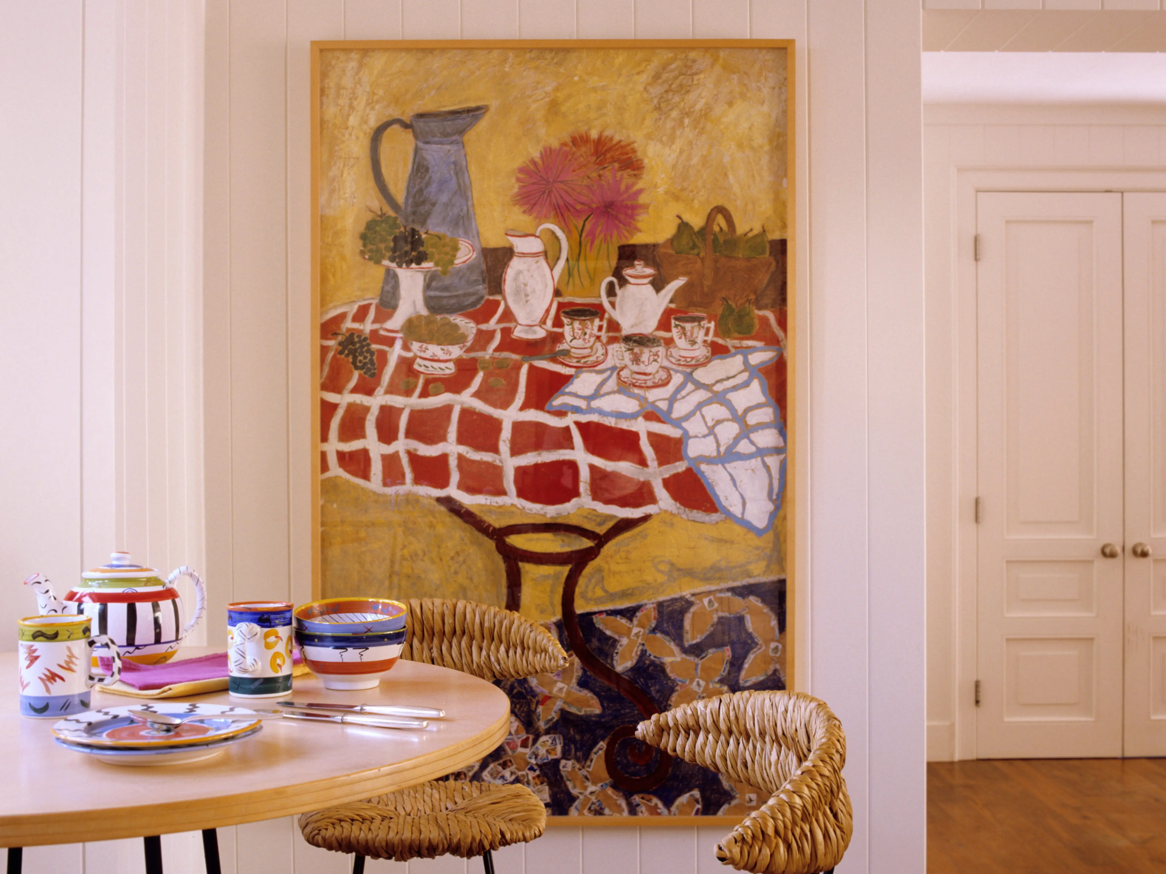 A room with a dining table with colorful dishes and a large painting of a dining table with teapots and flowers.