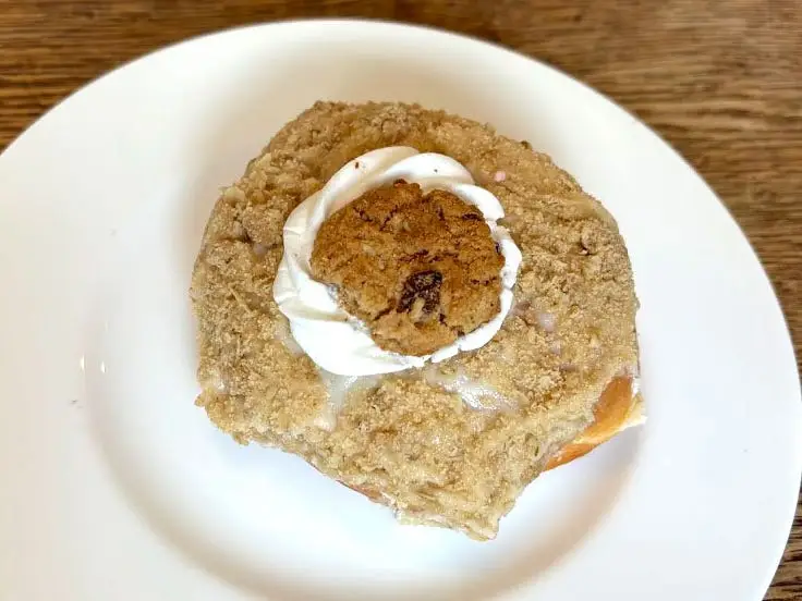 A doughnut topped with vanilla frosting, cinnamon streusel, a dollop of cream, and a small oatmeal cookie on a white plate.