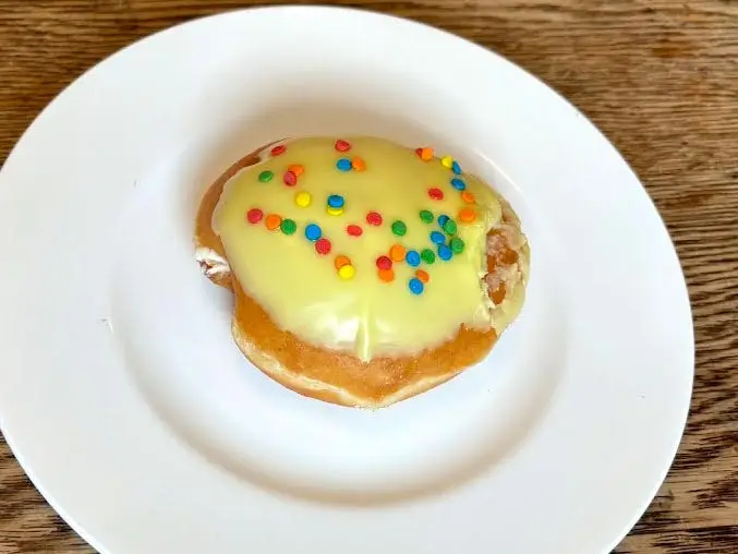 A doughnut with no hole that's topped with yellow icing and colorful sprinkles on a white plate.