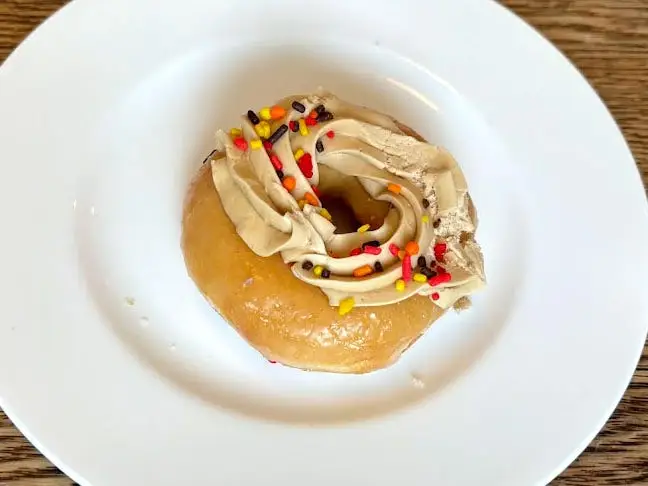 A glazed doughnut topped with thick beige frosting and colorful sprinkles on a white plate.