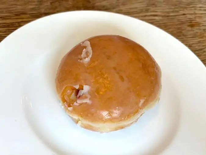 A glazed doughnut with no hole that's filled with raspberry jelly on a white plate.