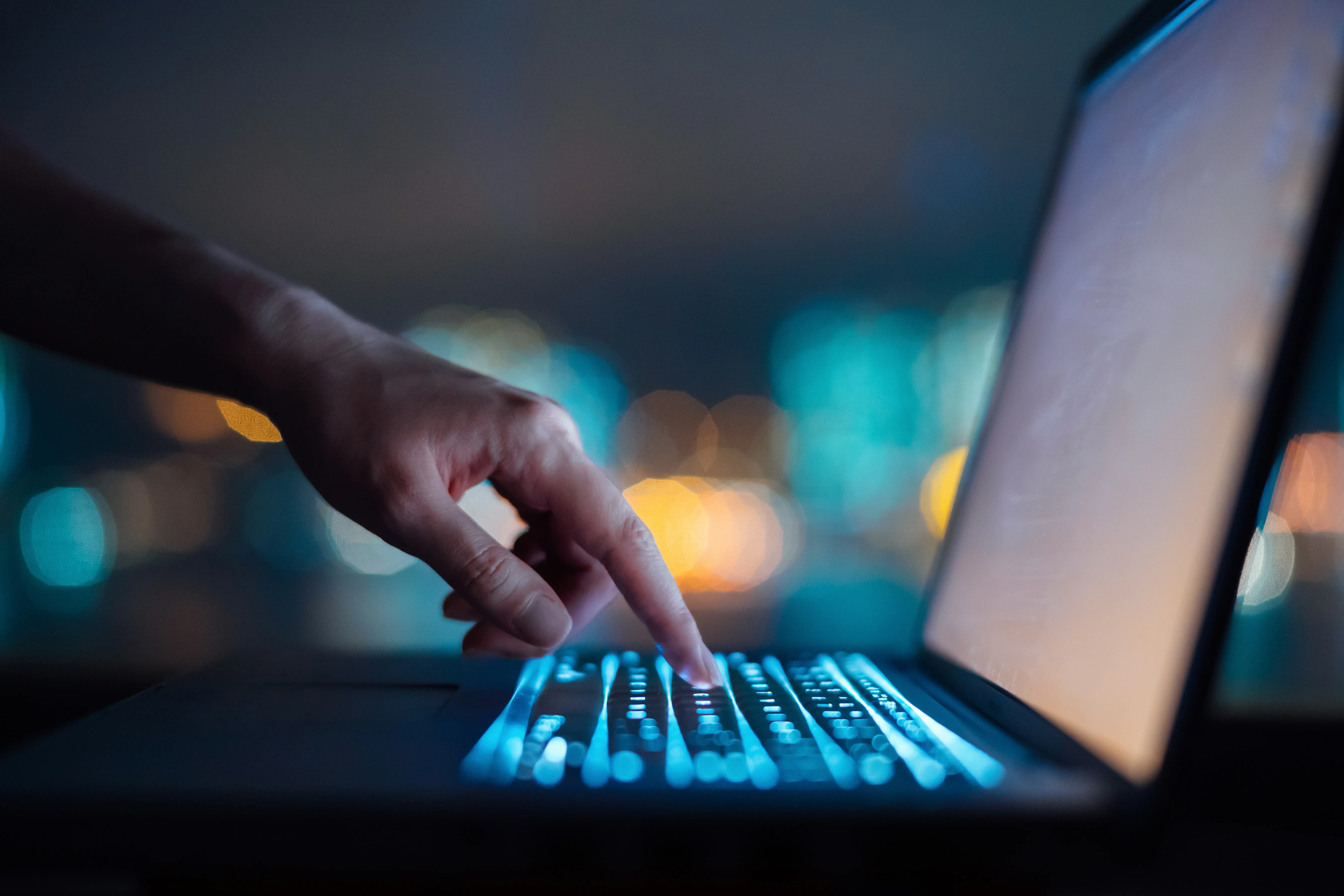 A person's finger types on a lit-up keyboard on their laptop.
