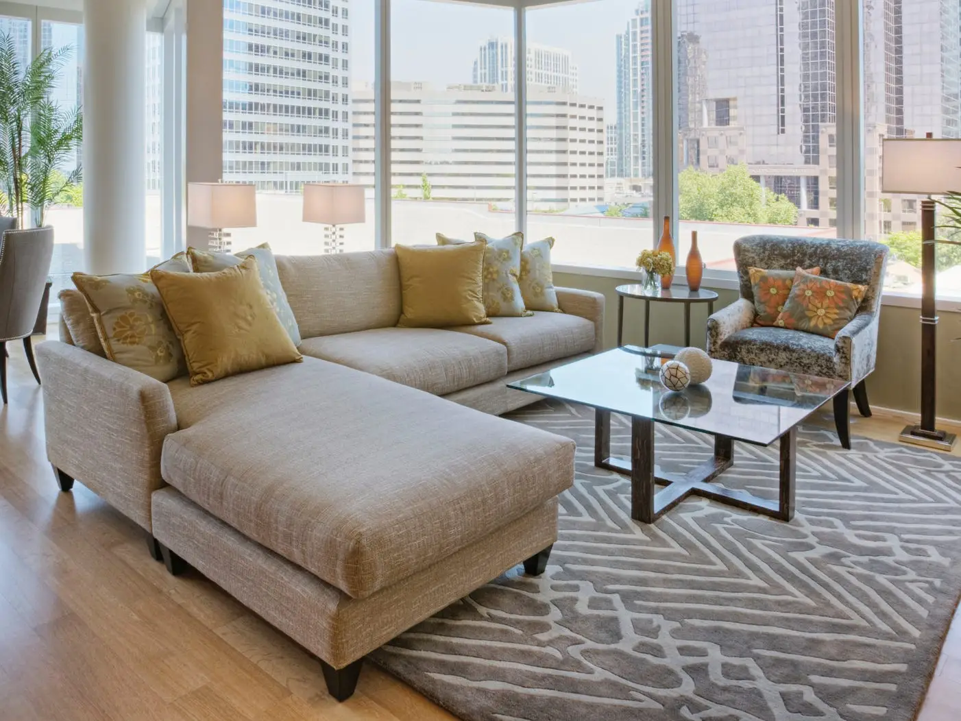 A living room with light wooden flooring and a carpet with a tan and white design with a beige sectional with loose back cushions and yellow pillows