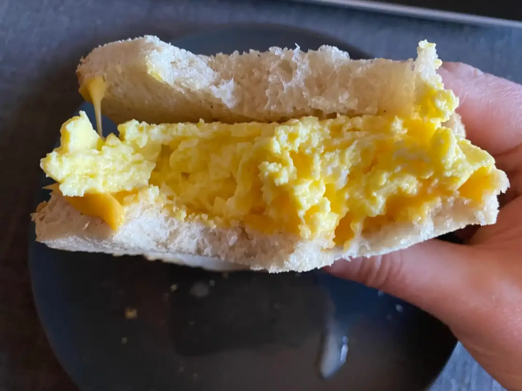 A hand holds a scrambled-egg sandwich over a dark-blue plate