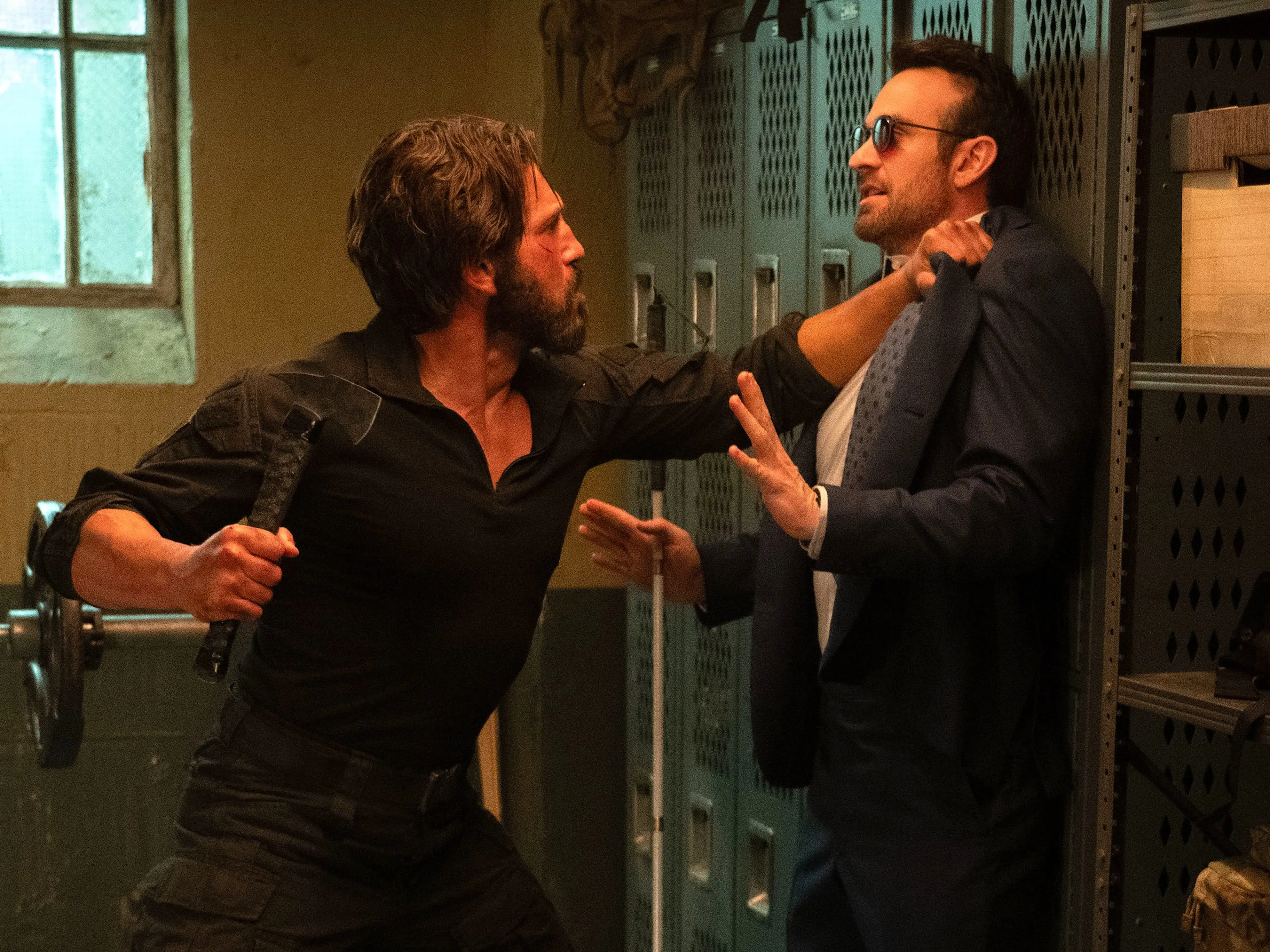 Two men standing in a locker room. On the left, a man in all-black military clothing has long black hair and a long black beard. He's holding a black hatchet in his right hand and he's holding the other man up against the lockers in the other. The other man is dressd in a dark blue suit and a white shirt. He's wearing dark glasses and he's holding a white cane in his left hand. There is a weight bench behind them, and tactical gear on shelves on the right.
