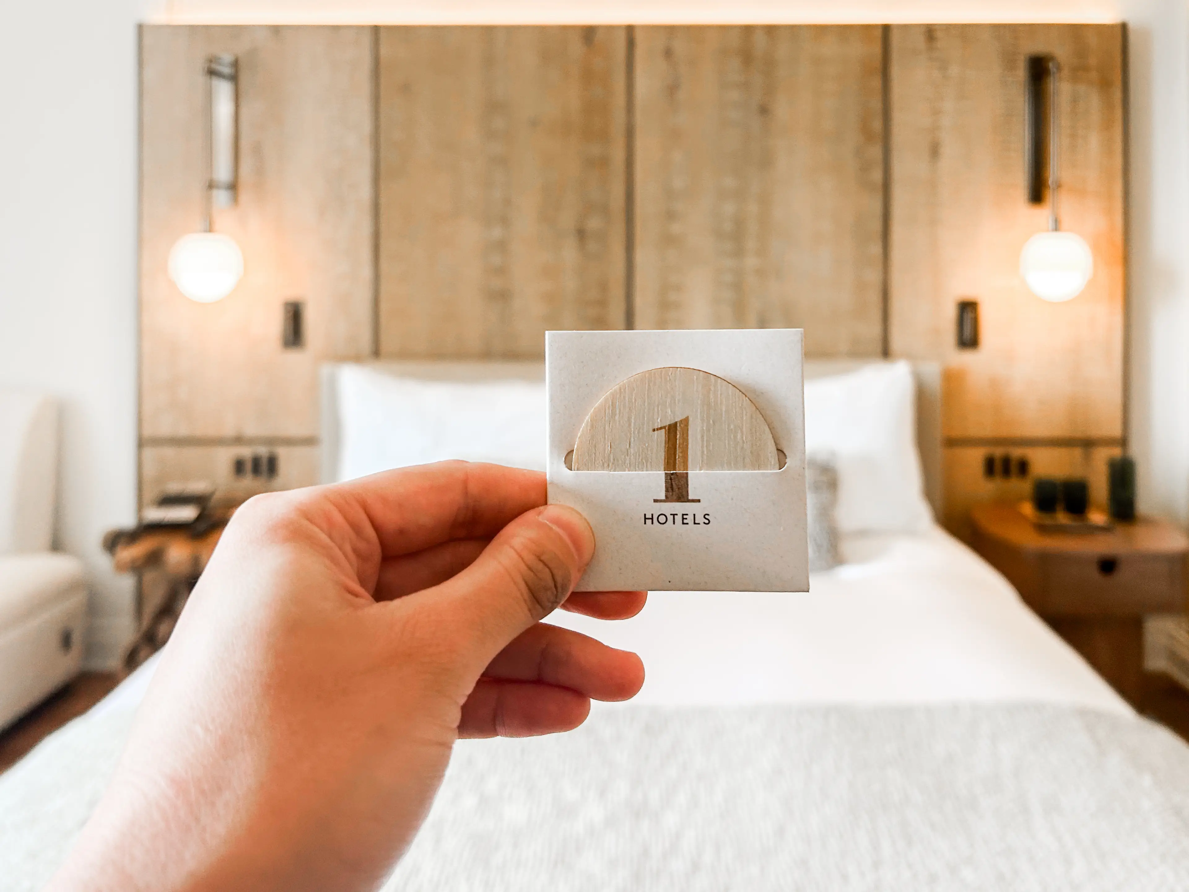A hand holds a round key card in front of a bed in a hotel room