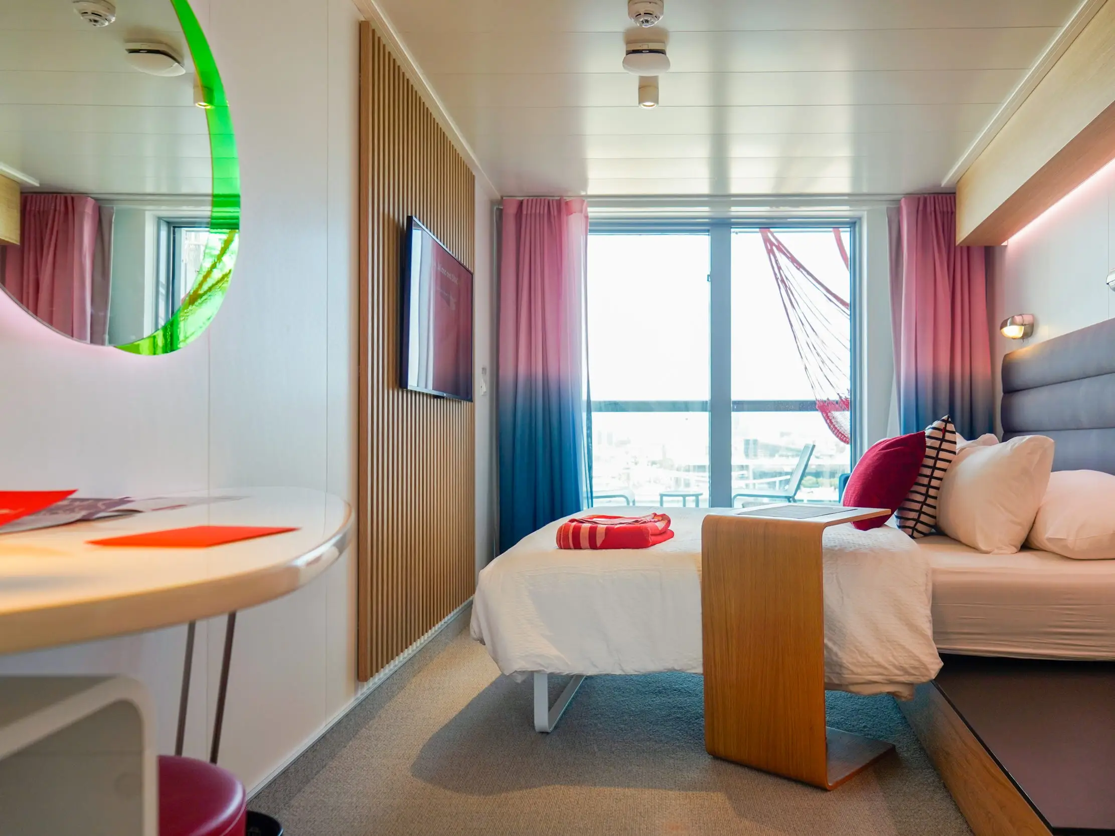A room with a mirror and table on the left. Behind the table is a bed with a navy headboard, white sheets, and white and red pillows as well as a red bolded blanket at the bottom seen from the side in front of a large window with red and blue curtains. There's a mountain TV on a wooden-planed wall to the left  and a wooden side table next to the bed.