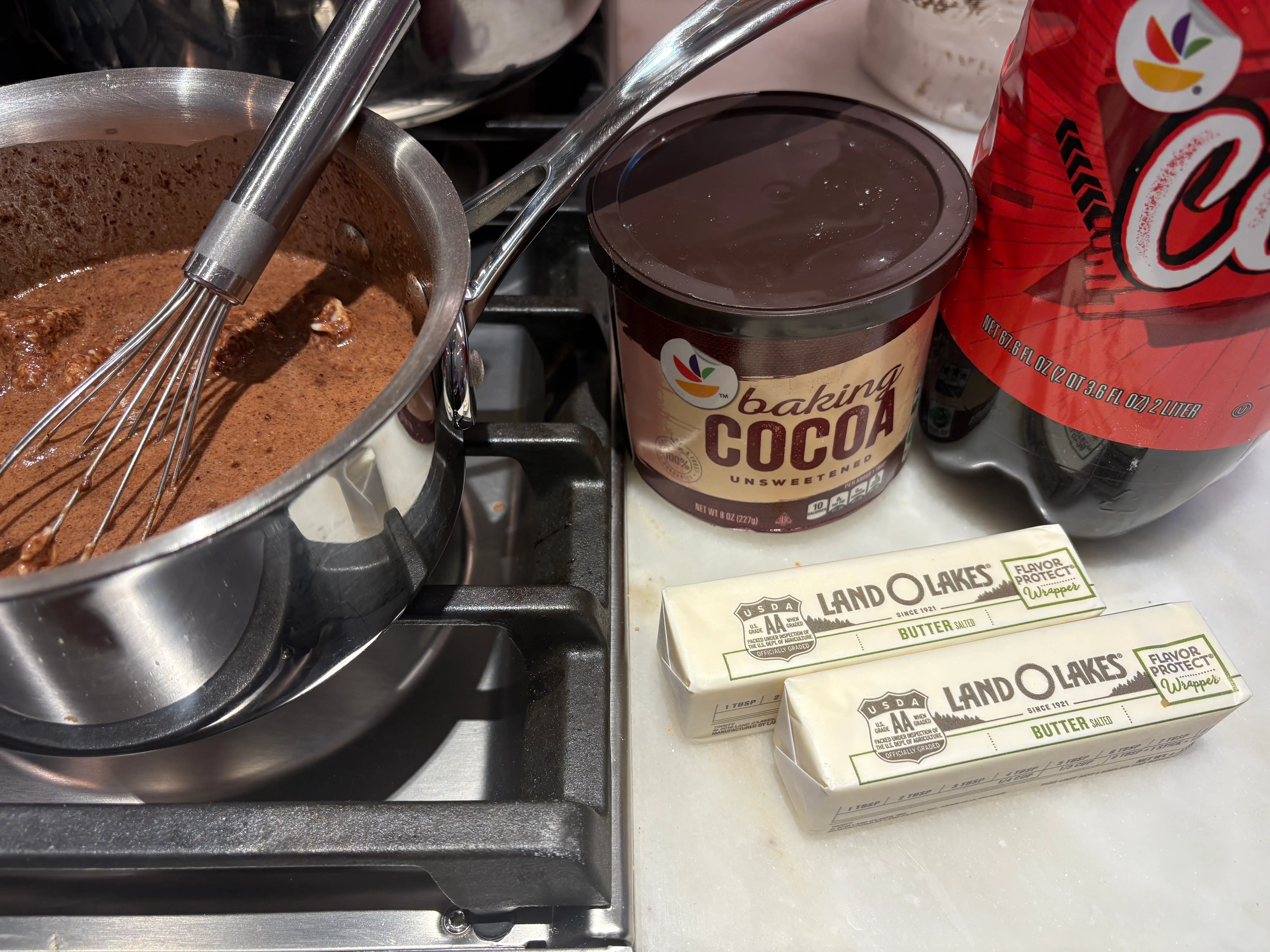 The butter, cocoa powder, and cola on the counter with the mixture on the stove.