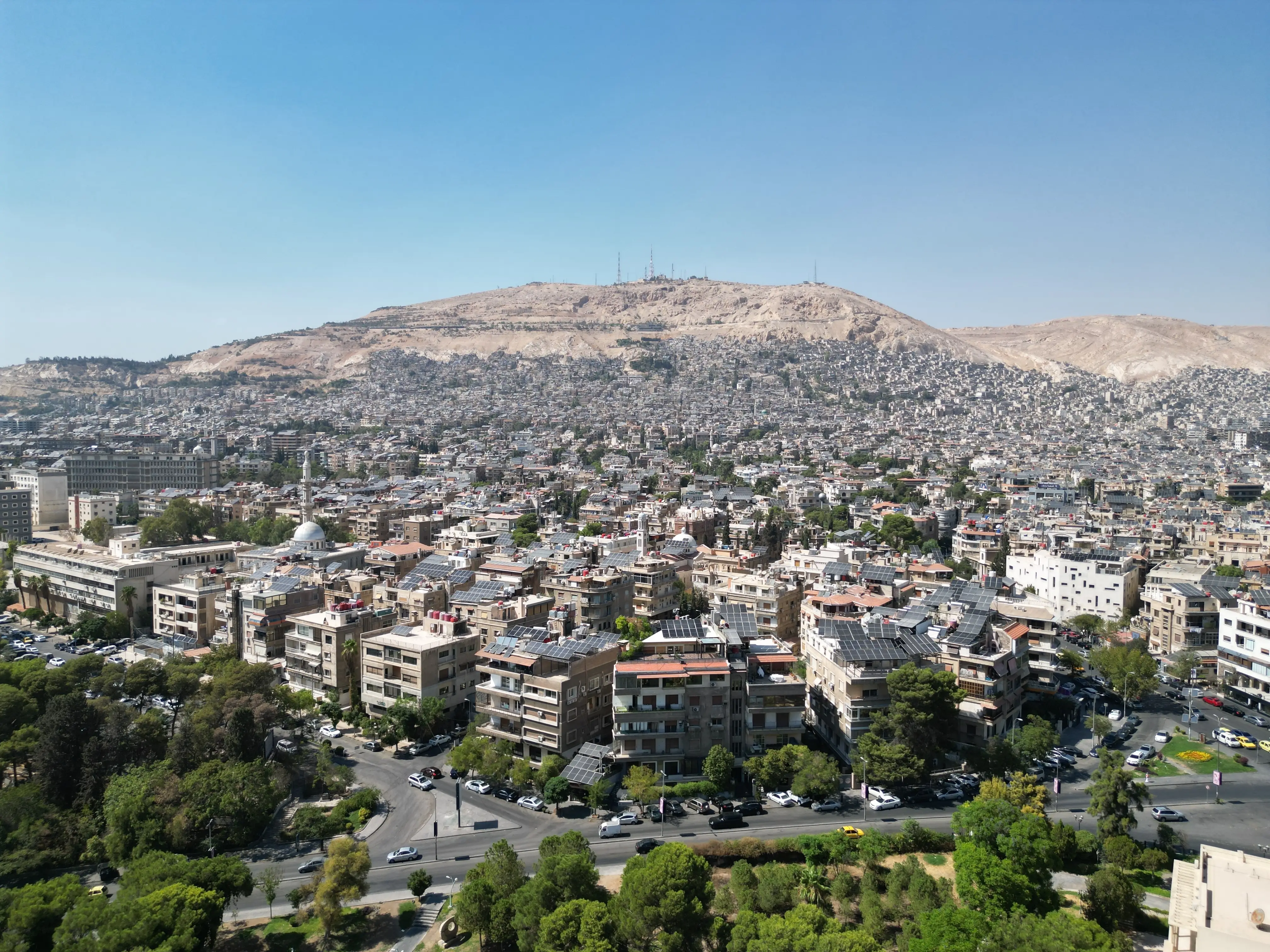An aerial view captures the new Syrian national flag hoisted on a towering pole in central Damascus, with Mount Qasioun, one of the capital's most prominent landmarks rising in the background.