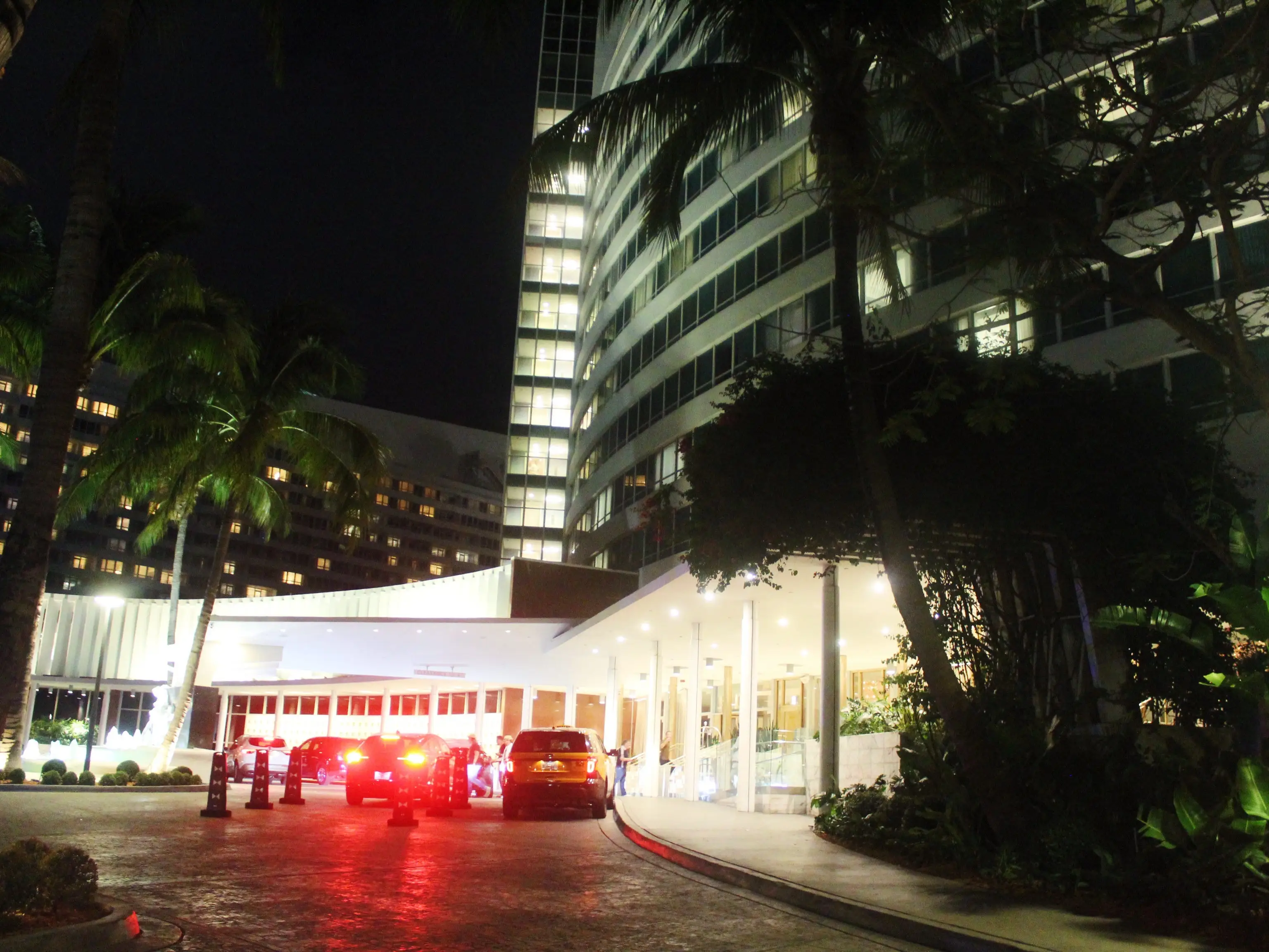 Cars with glowing taillights wait beneath a brightly lit high-rise entrance lined with palm trees at night.