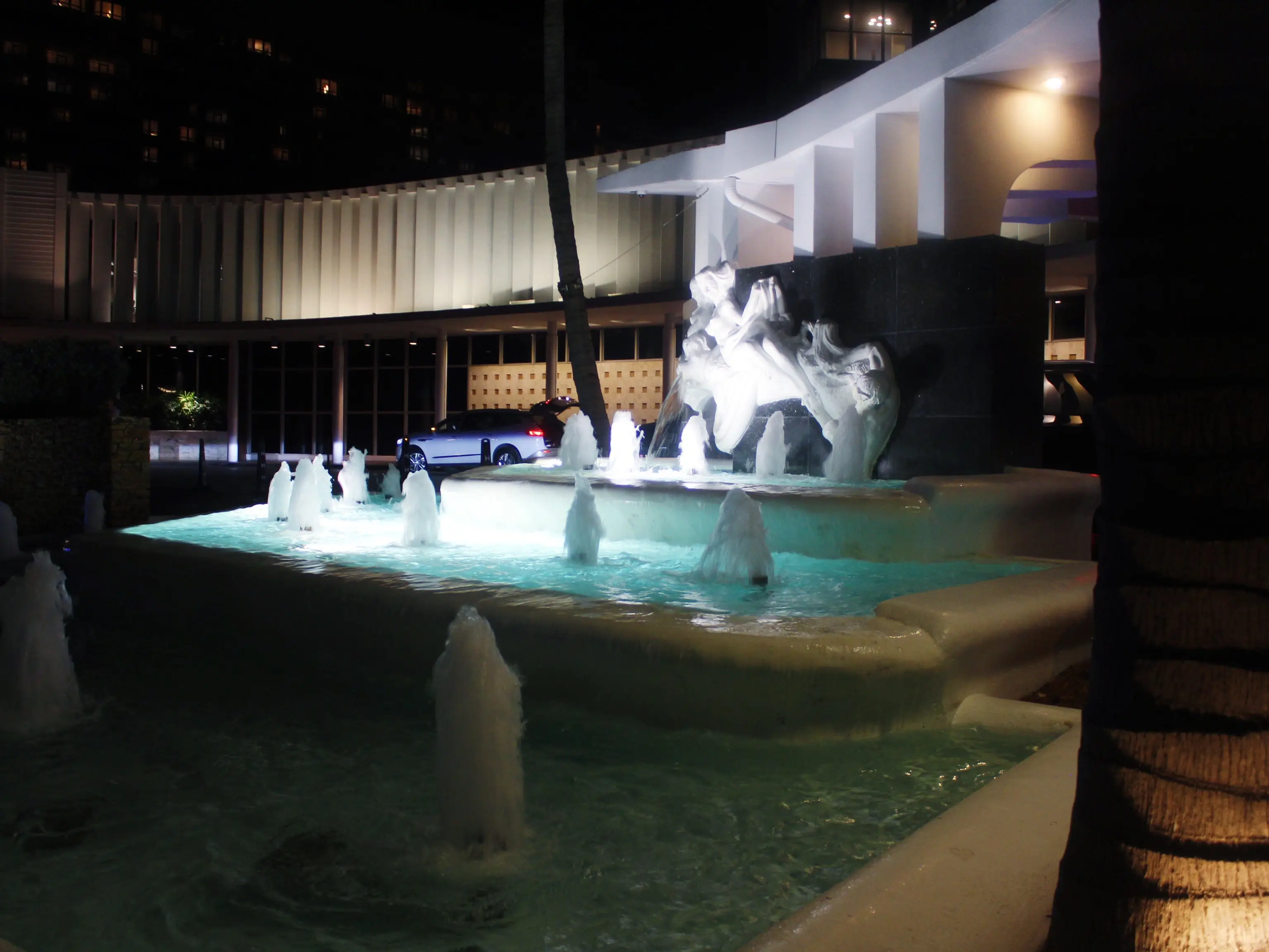 Illuminated outdoor fountain with sculptural water feature and palm trees outside a modern building at night.