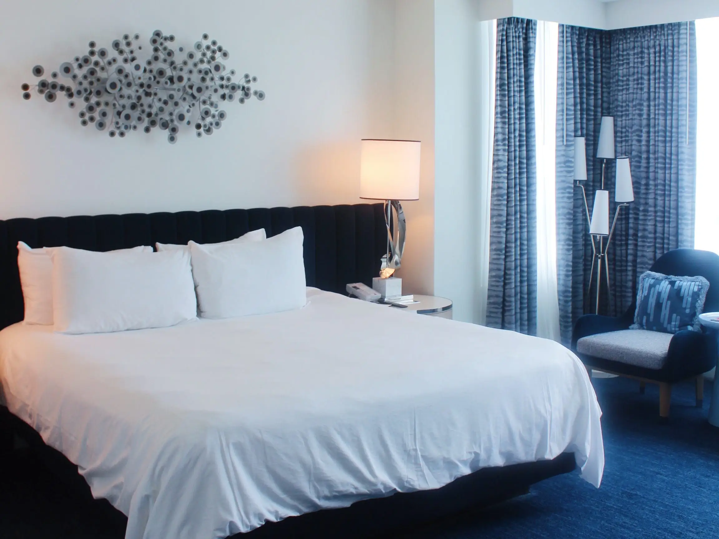 Modern hotel bedroom with a large white bed, blue carpet, wall art, lamps, curtains, and a lounge chair.