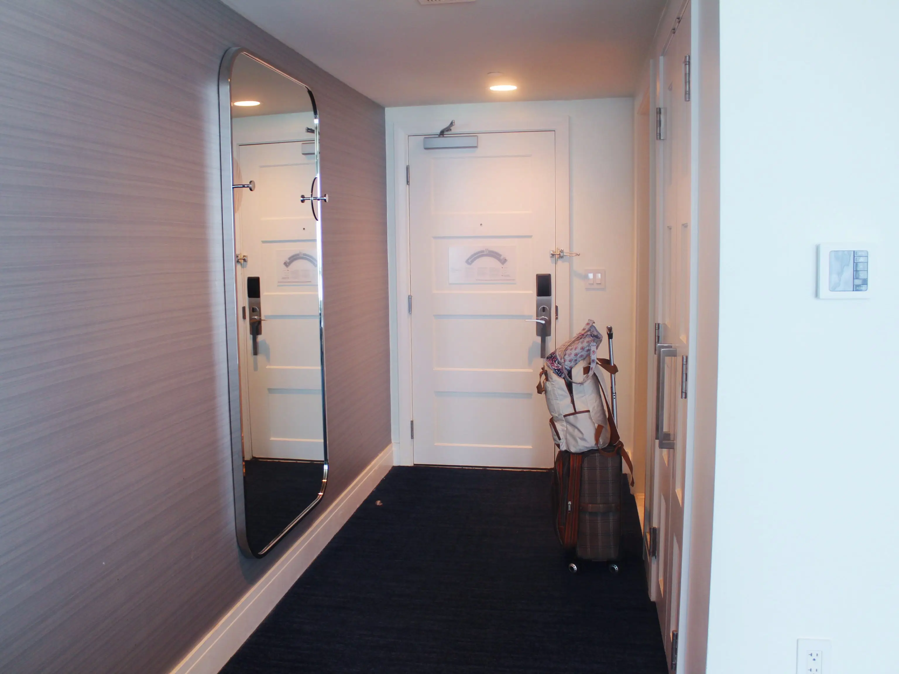 Hotel room entryway with a white door, wall mirror, dark carpet, and luggage standing near the entrance.