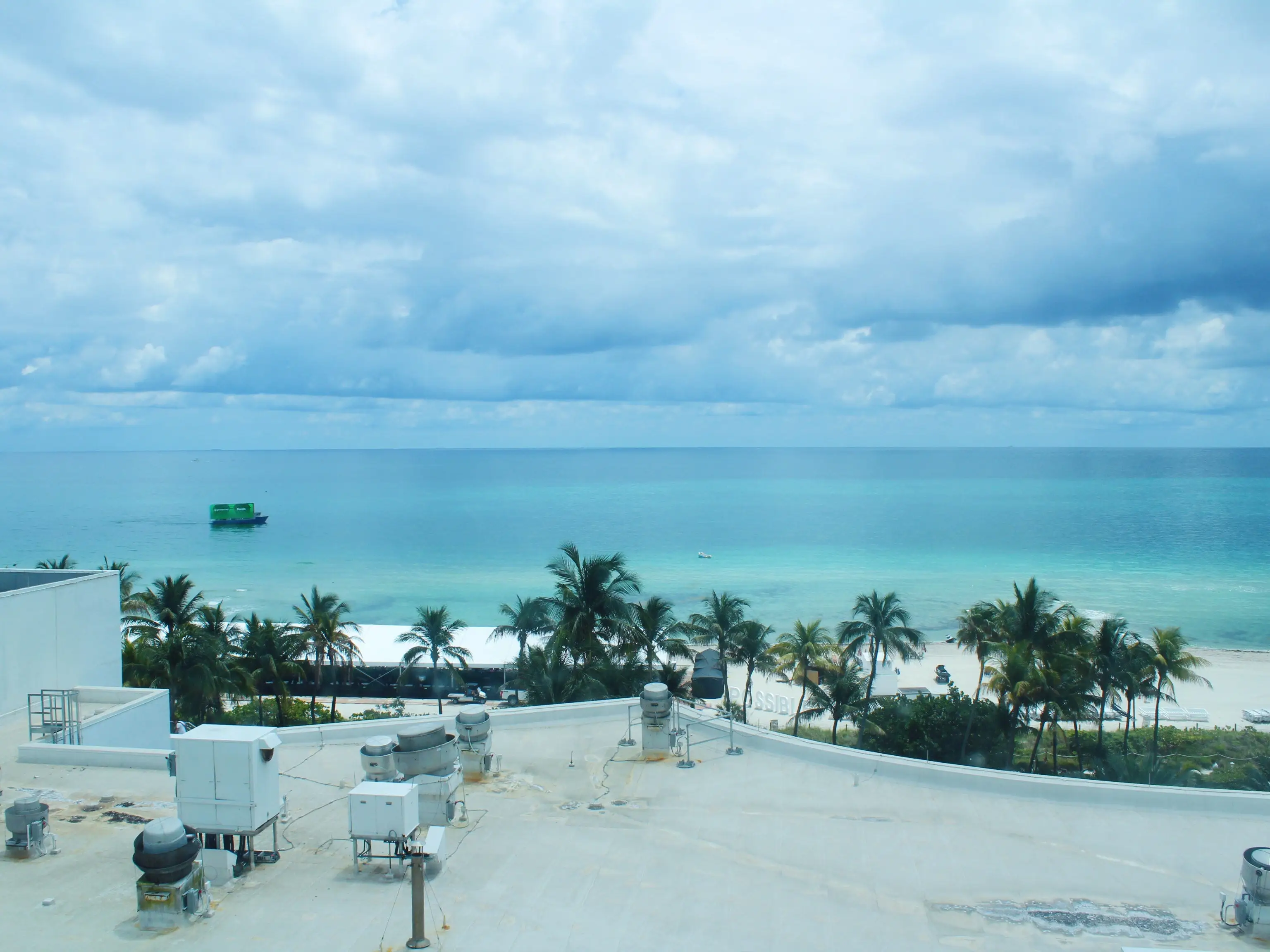 Rooftop equipment overlooks palm trees, a white sand beach, turquoise ocean water, and a cloudy sky.