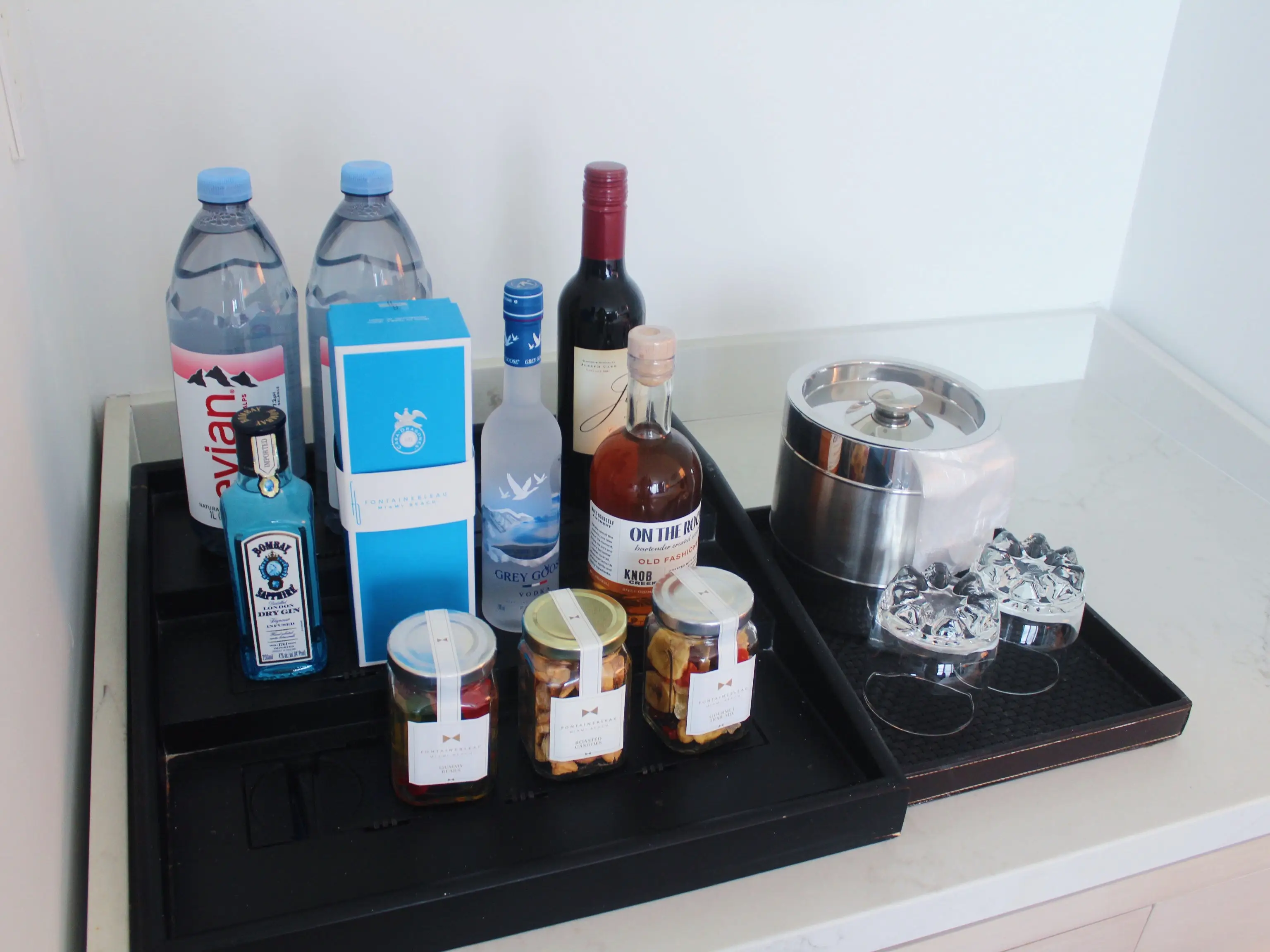 Hotel minibar tray displays assorted bottled water, spirits, snack jars, glasses, and a metal ice bucket.