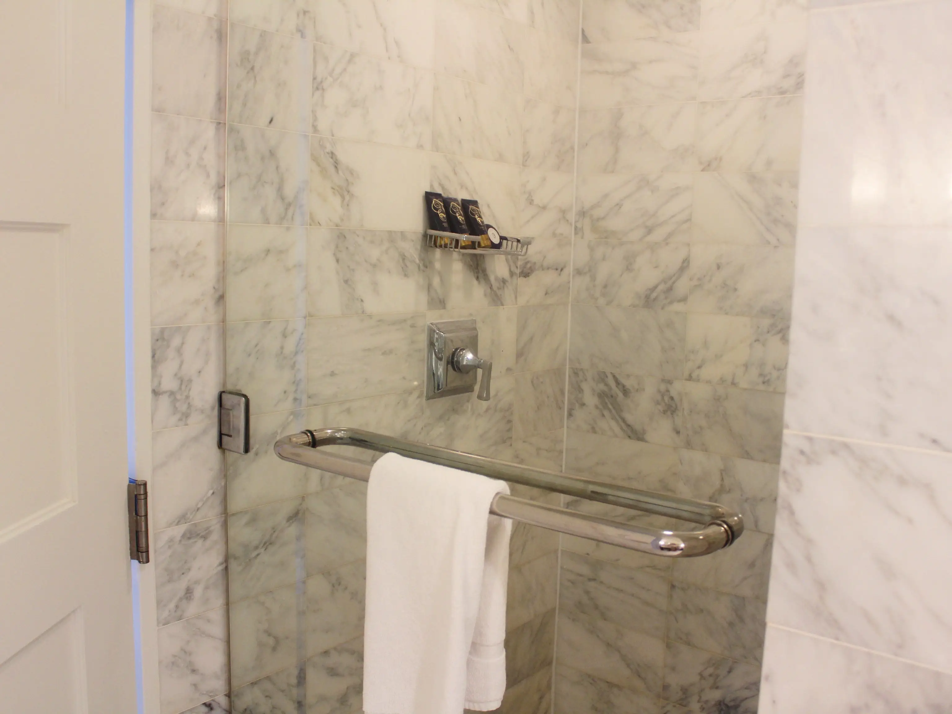 Marble-tiled shower with a glass door, chrome grab bar, white towel, wall controls, and small toiletries on a shelf.