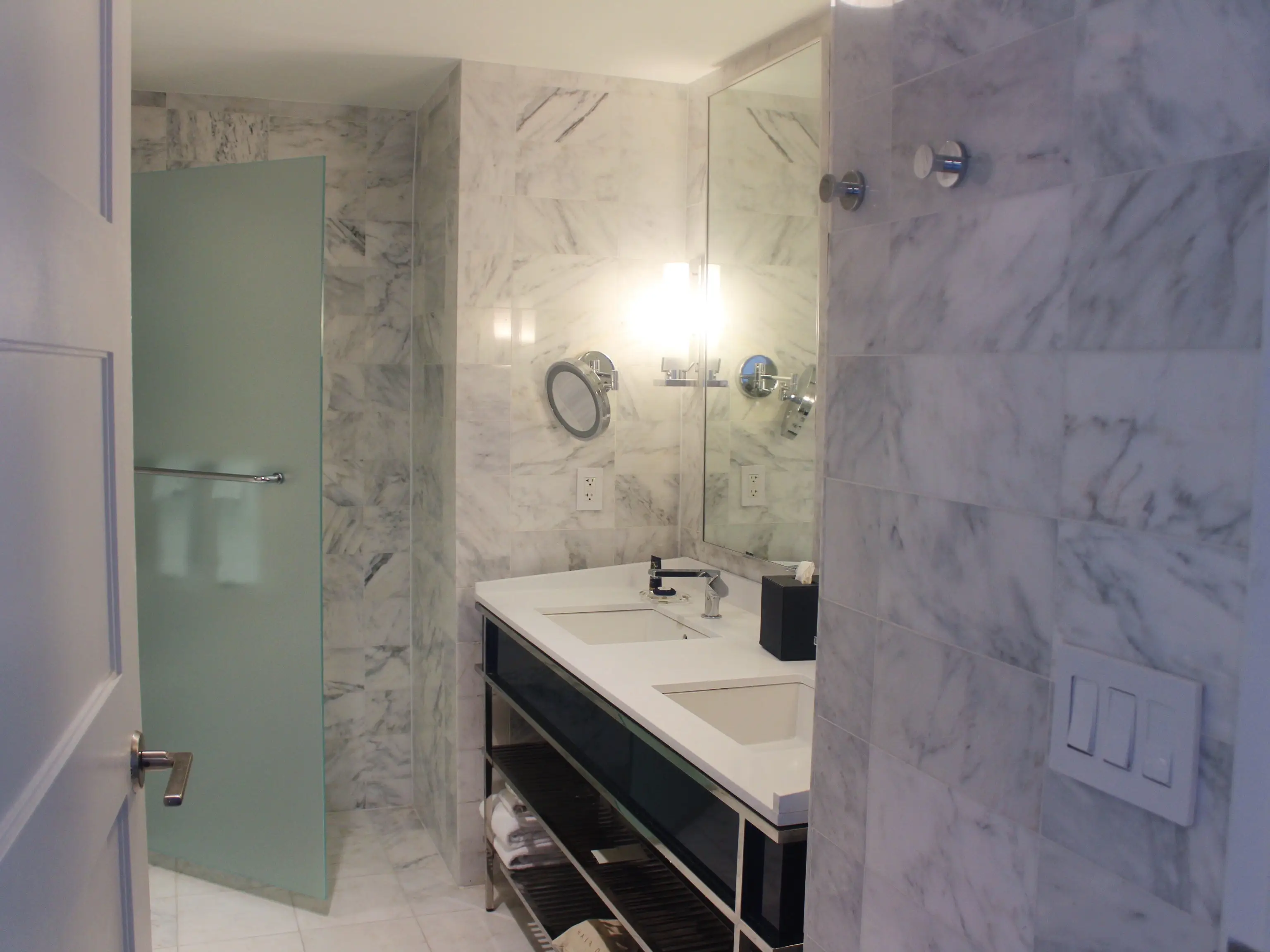 Modern marble bathroom with double sink vanity, large mirror, wall lights, and frosted glass shower door.