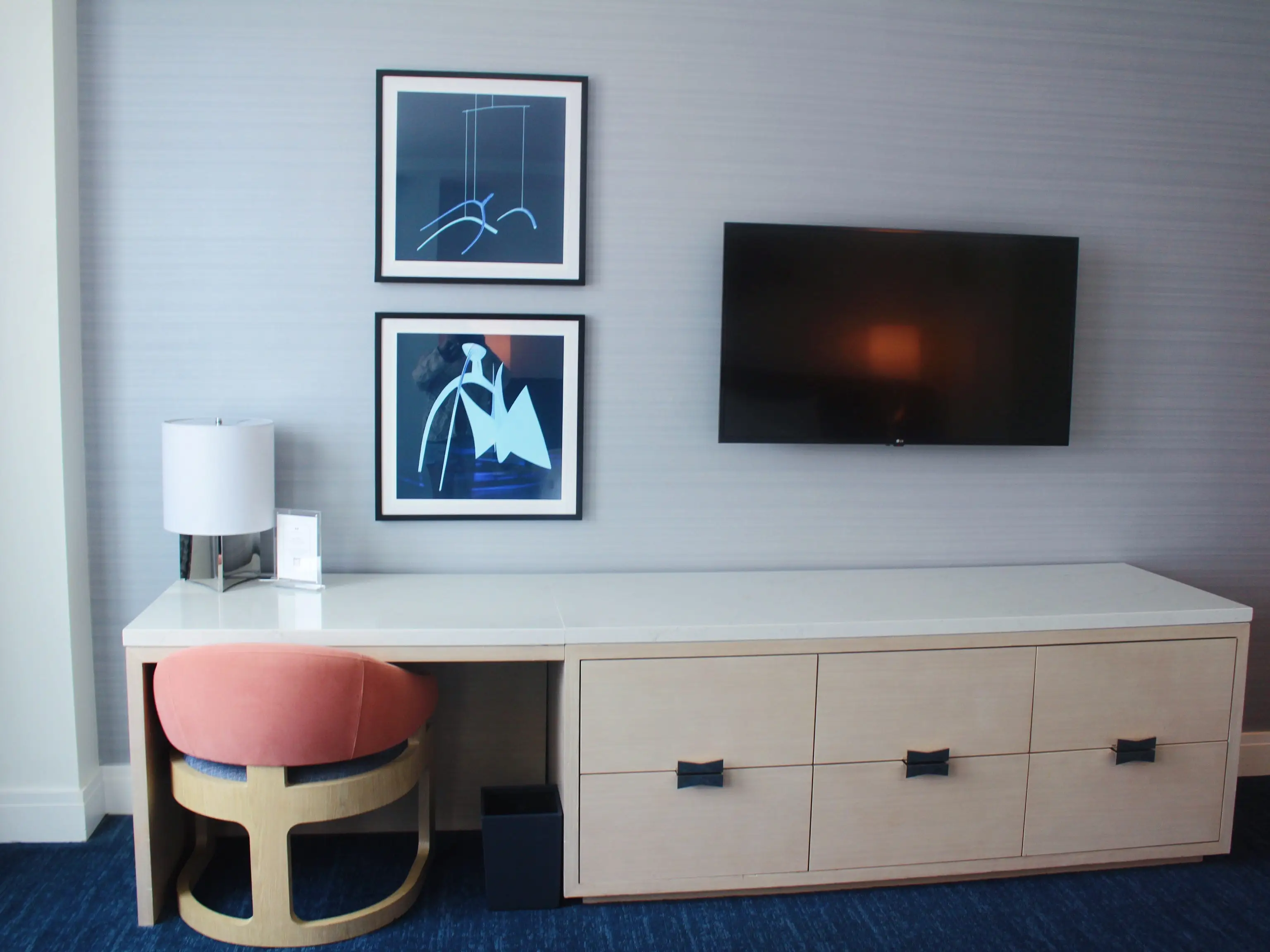 Modern room workspace with a wall-mounted television, framed abstract art, desk lamp, chair, and light wood dresser.